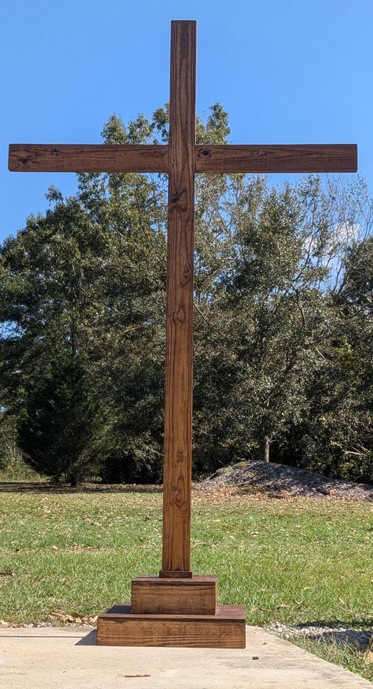 Wood cross outdoors against a backdrop of trees and a bright blue sky, featuring a rich brown stain, visible wood grain, and a three-tiered base.