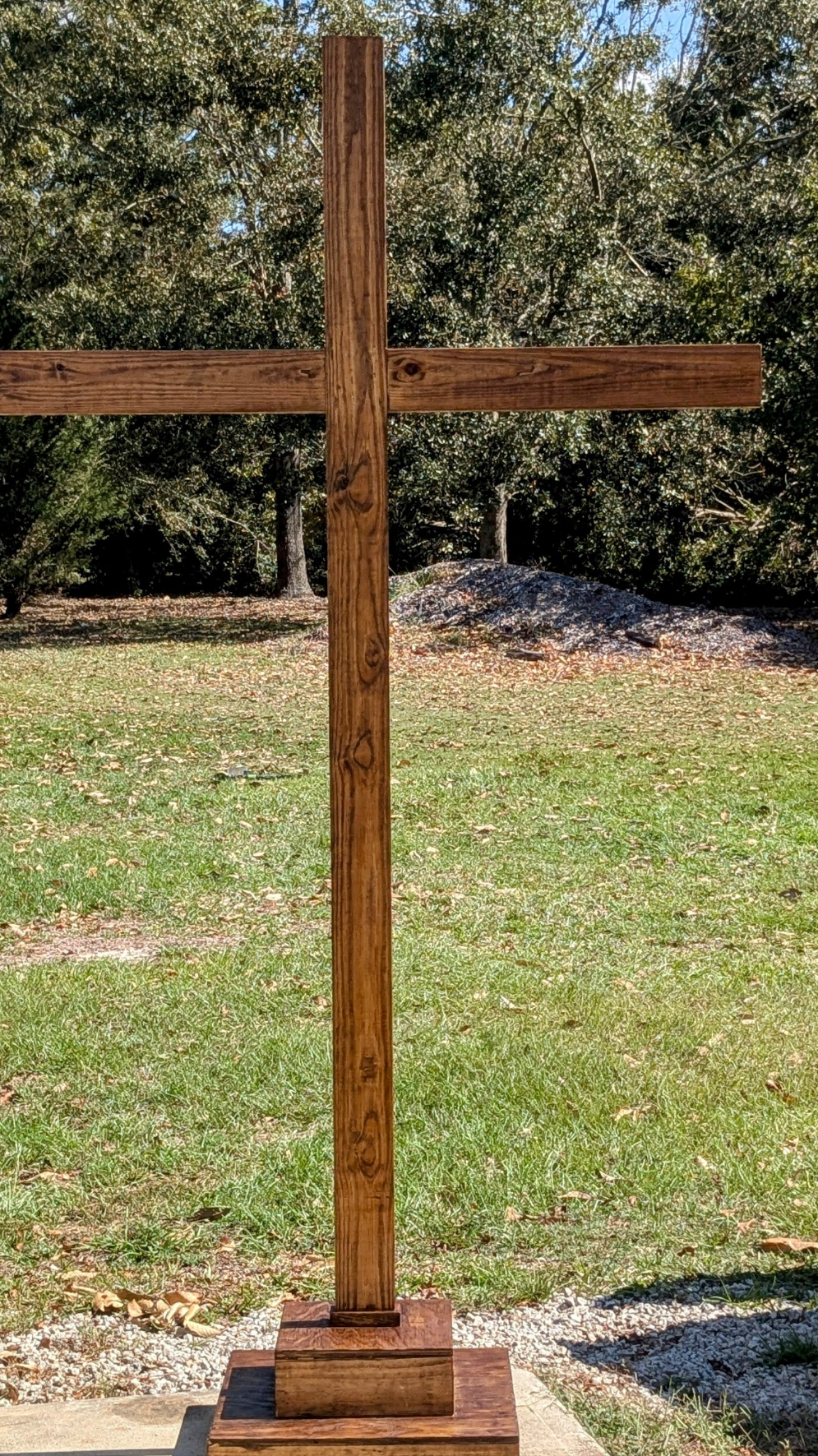Wooden cross outdoors in a grassy yard with trees in the background showing rich brown stain and visible wood grain
