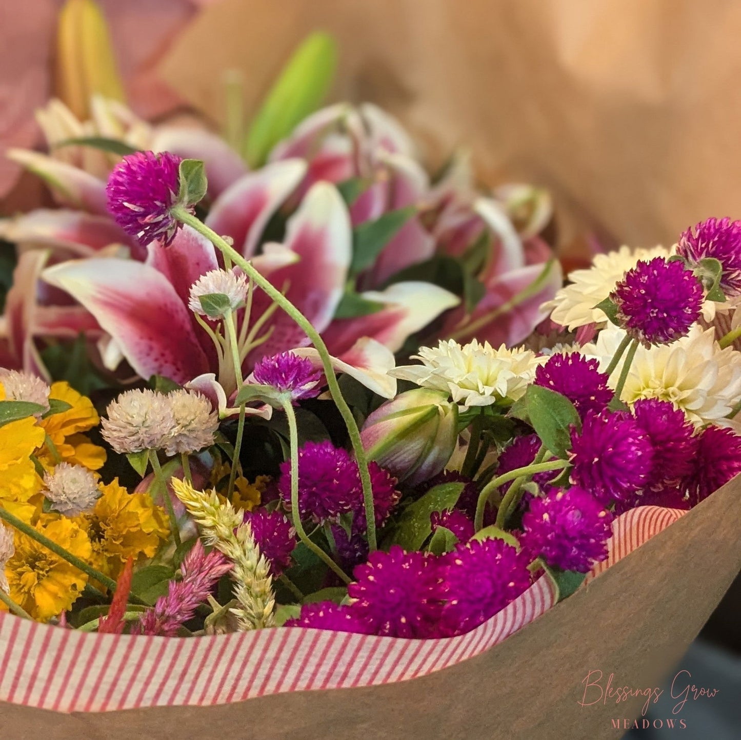 Blessings Grow Meadows bouquet featuring vibrant pink lilies, bright yellow marigolds, and deep purple gomphrena globosa flowers wrapped in pink and white striped paper.
