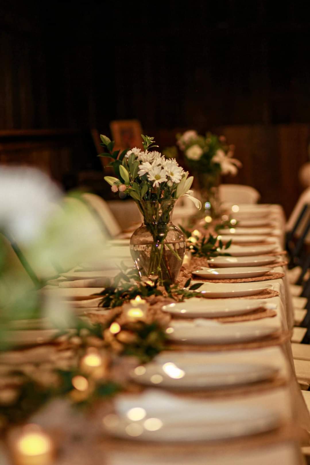 Photograph of a rustic wedding reception tablescape featuring white daisies and lilies in a clear glass vase with candlelight and natural greenery accents along a long table