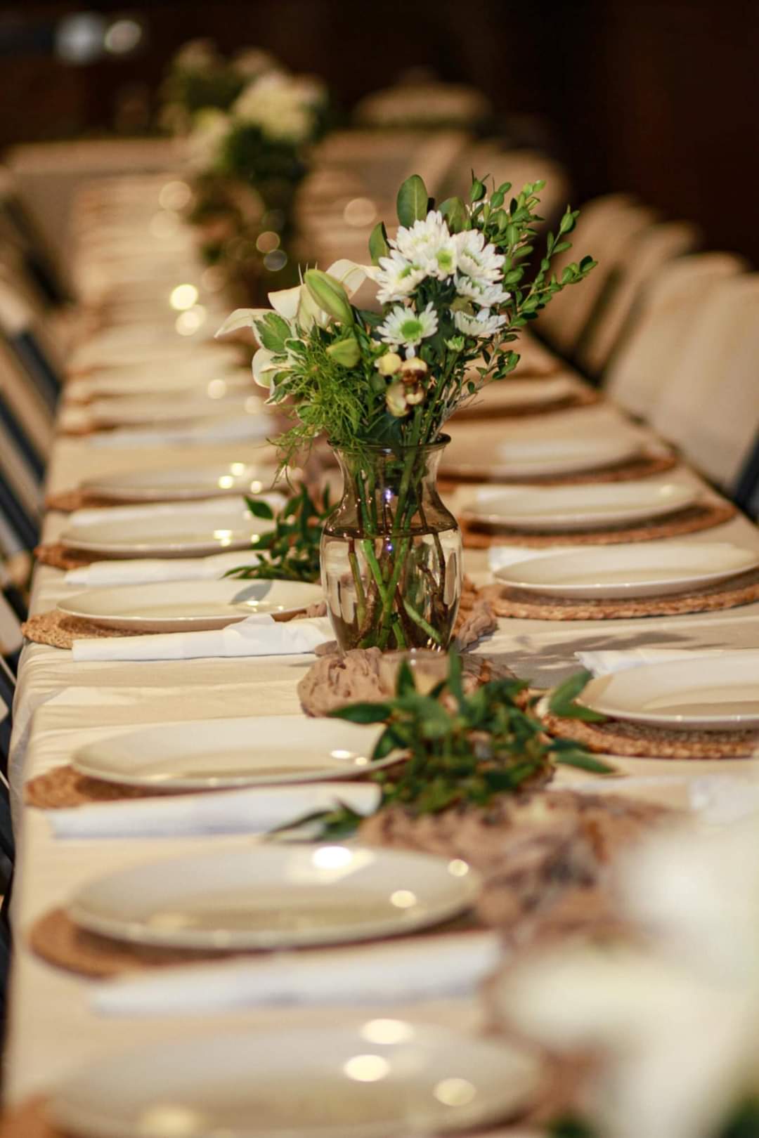 Photography wedding reception centerpiece featuring white daisies lilies and greenery in a glass vase on a long table with white plates and woven placemats