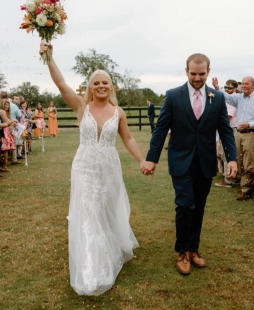 Photograph of a bride in a white lace gown and groom in a navy suit walking down a grassy aisle past a wooden fence with a colorful bouquet and flower petals a rustic outdoor wedding venue

