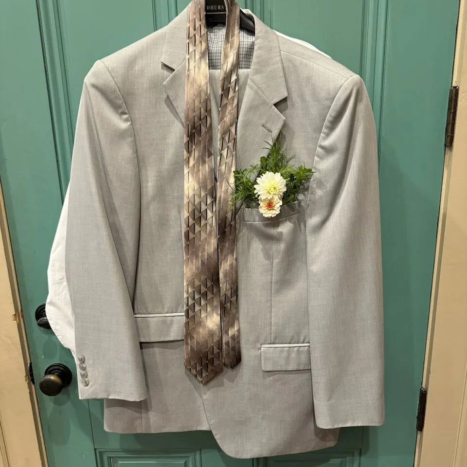 A light gray suit jacket hangs on a teal door with two patterned neckties around the collar and a small white boutonniere made from Blessings Grow Meadows Merch & More DIY Event Flowers + Bulk Fresh Cut Greenery pinned to the left lapel.