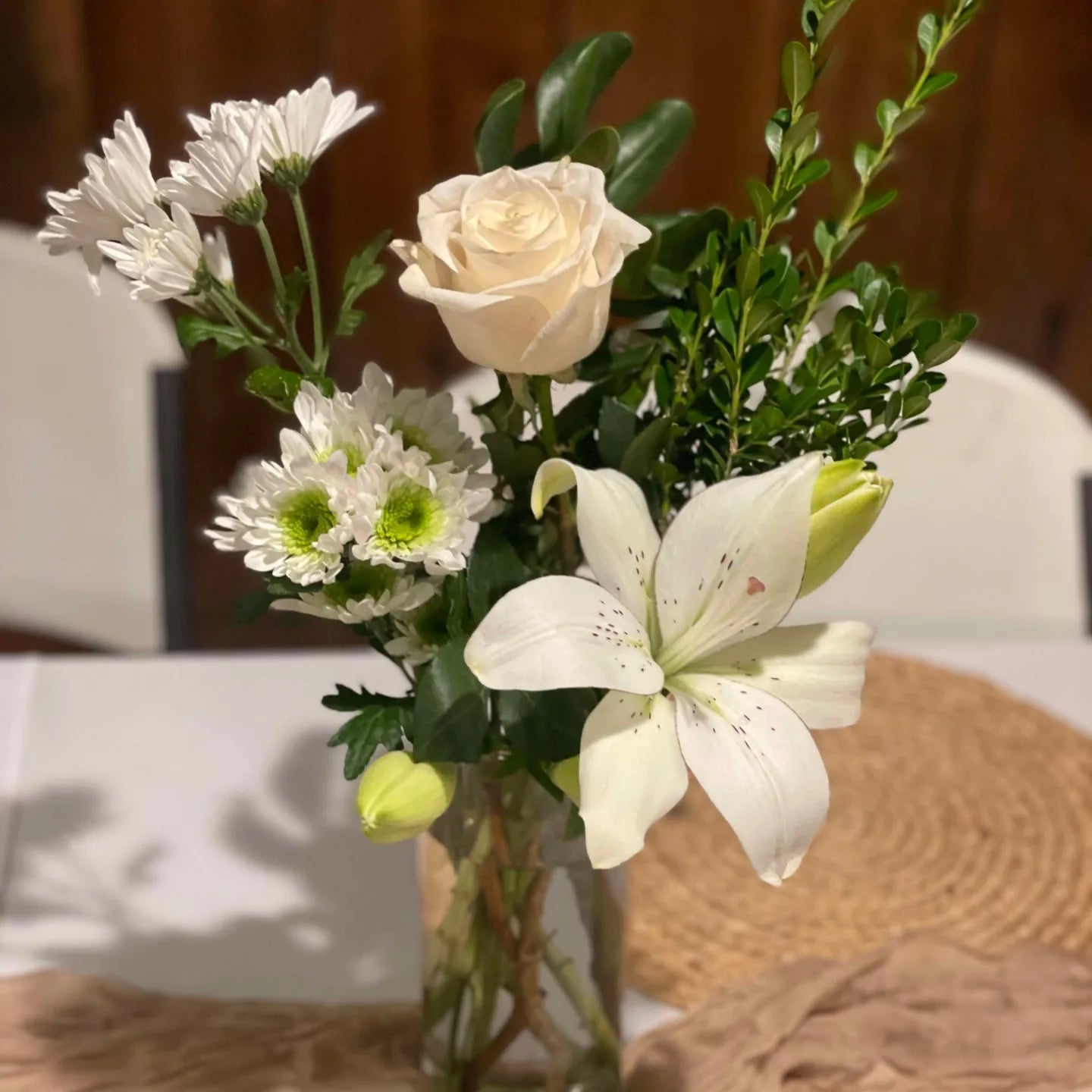 Photograph of a table arrangement featuring white daisies, a creamy rose, and a lily with hints of maroon in a glass vase on a beige placemat
