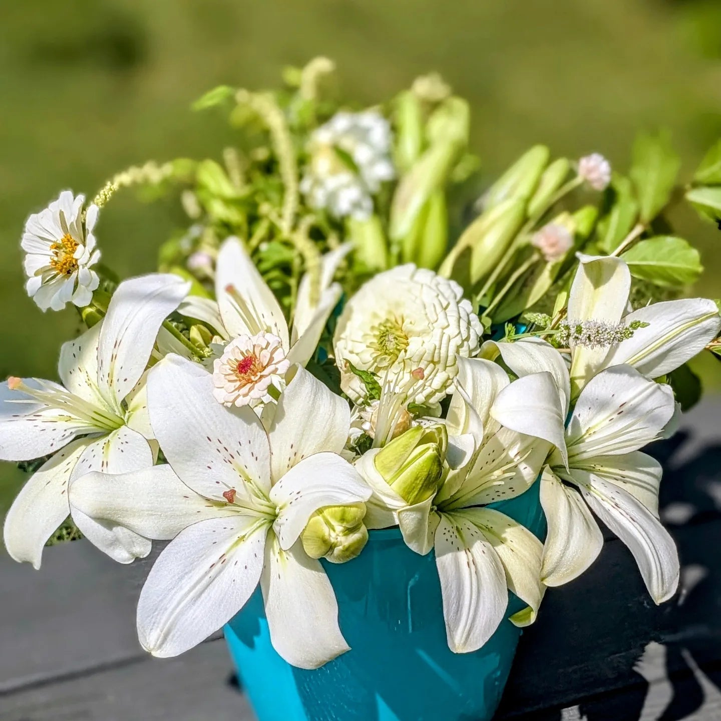 Photograph of a floral arrangement featuring prominent white lilies, zinnias, and dahlias in a teal bucket outdoors