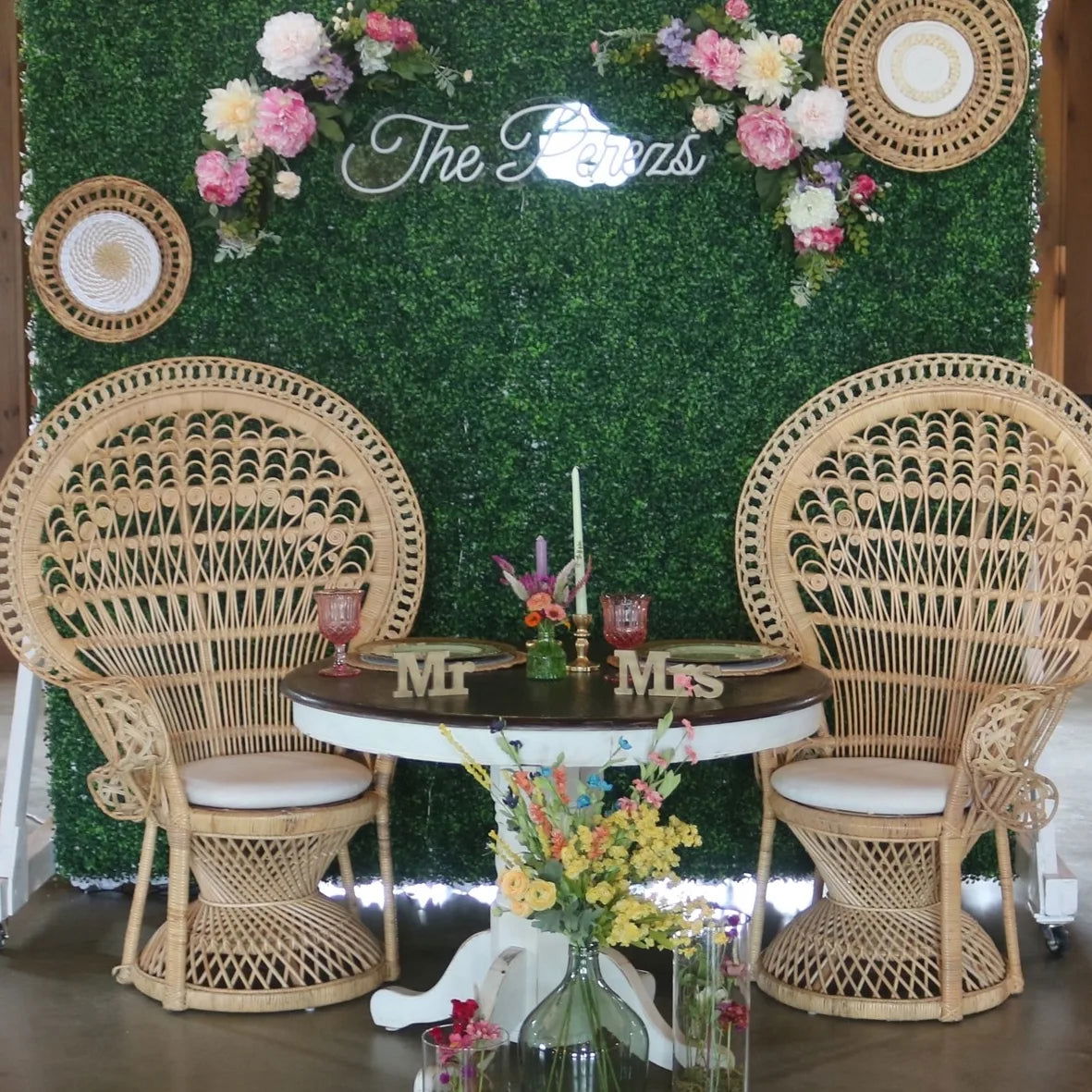 A romantic table for two with wicker chairs, a round table, pink goblets, and “Mr & Mrs” signs is set against a green leafy backdrop with fresh DIY Event Flowers + Bulk Greenery from Blessings Grow Meadows Merch & More’s Bloom Buckets (Metter, GA pick-up).