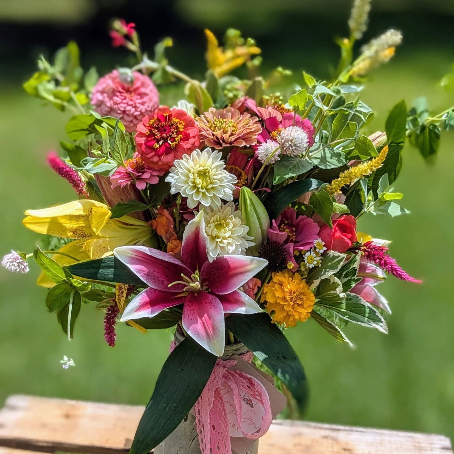 The Custom Deluxe Arrangement by Blessings Grow Meadows Merch & More features assorted, locally grown pink lilies, zinnias, dahlias, and greenery in a jar with a pink ribbon, beautifully set outdoors against a blurred green background.