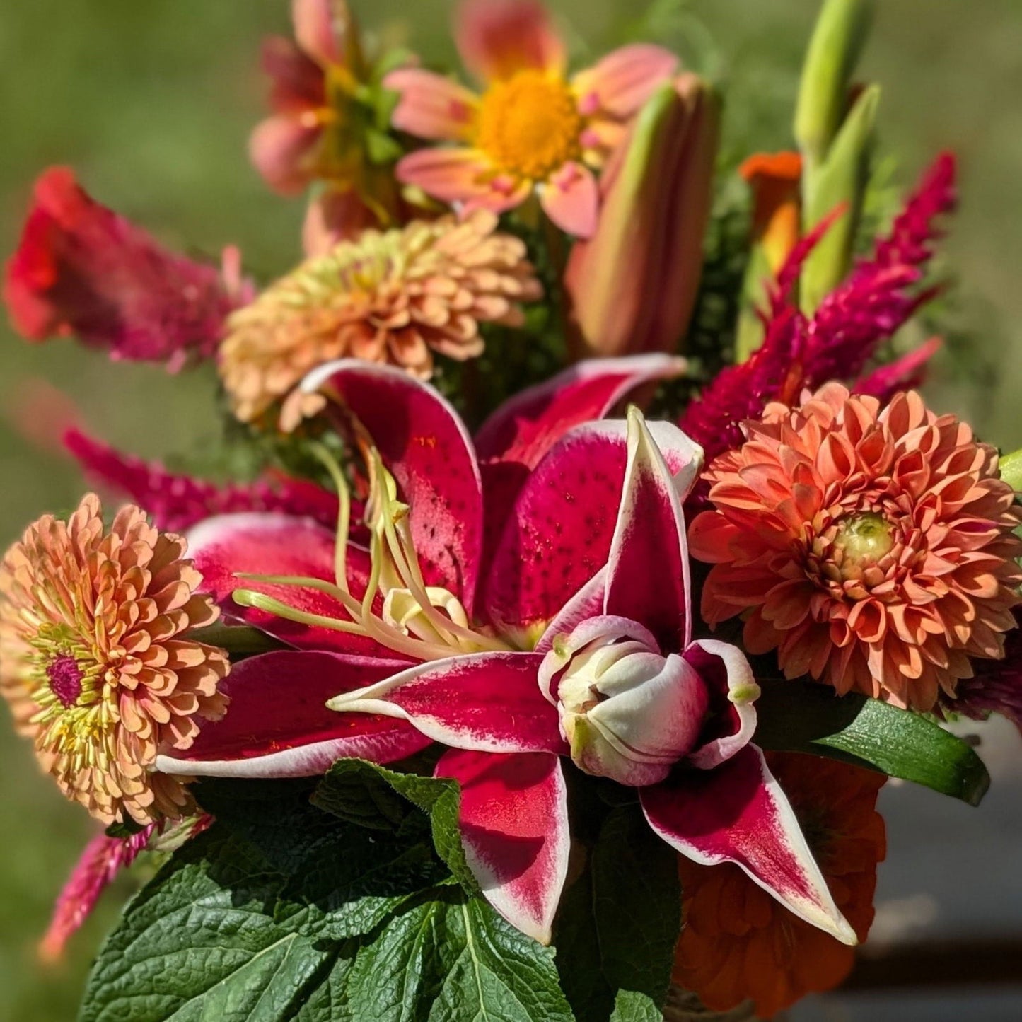 The Custom Deluxe Arrangement by Blessings Grow Meadows Merch & More is a vibrant bouquet of pink lilies, orange zinnias, and assorted locally grown flowers with green leaves, set against a blurred outdoor background.