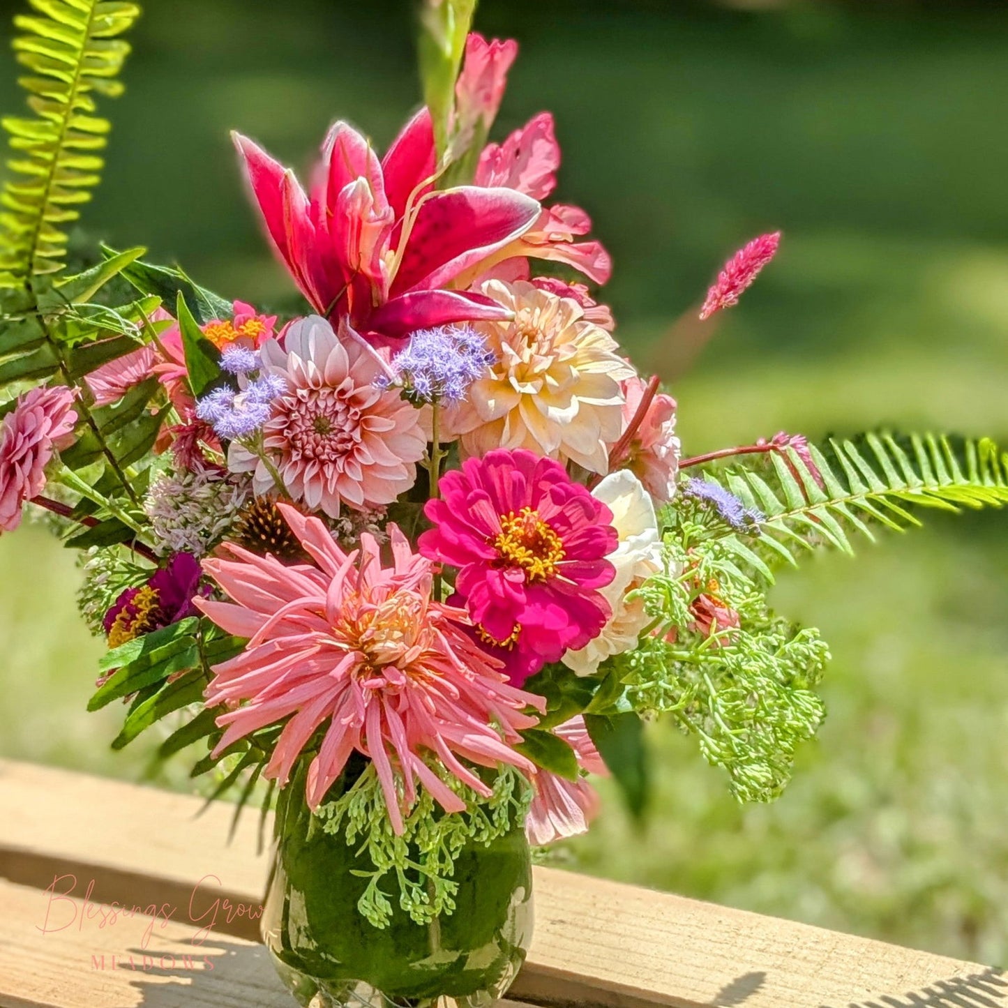 Blessings Grow Meadows floral arrangement featuring dahlias, zinnias, and lilies in a green glass vase outdoors with vibrant pink, bright fuchsia, and soft lavender tones.
