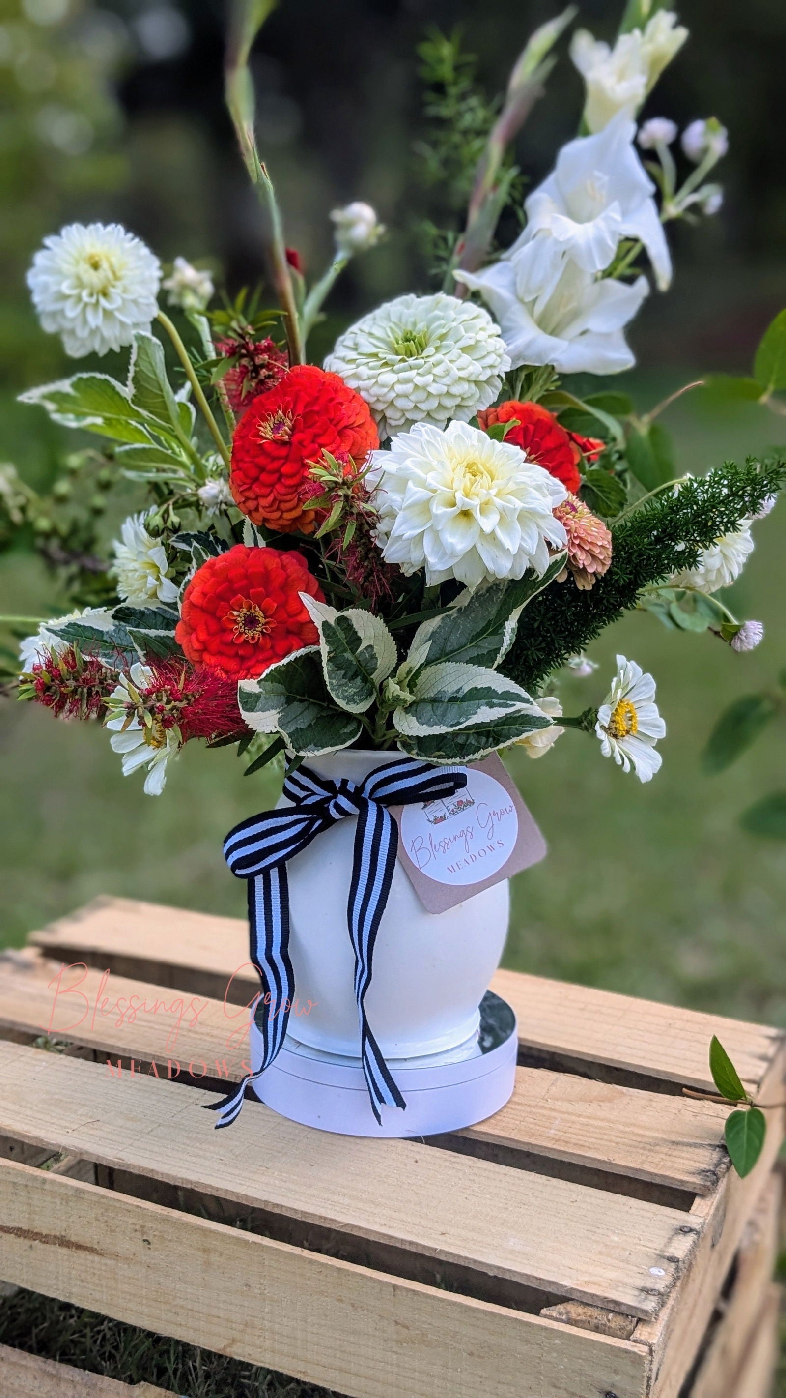 Blessings Grow Meadows floral arrangement featuring dahlias zinnias and gladioli in a white vase with a black and white ribbon on a rustic wooden crate outdoors
