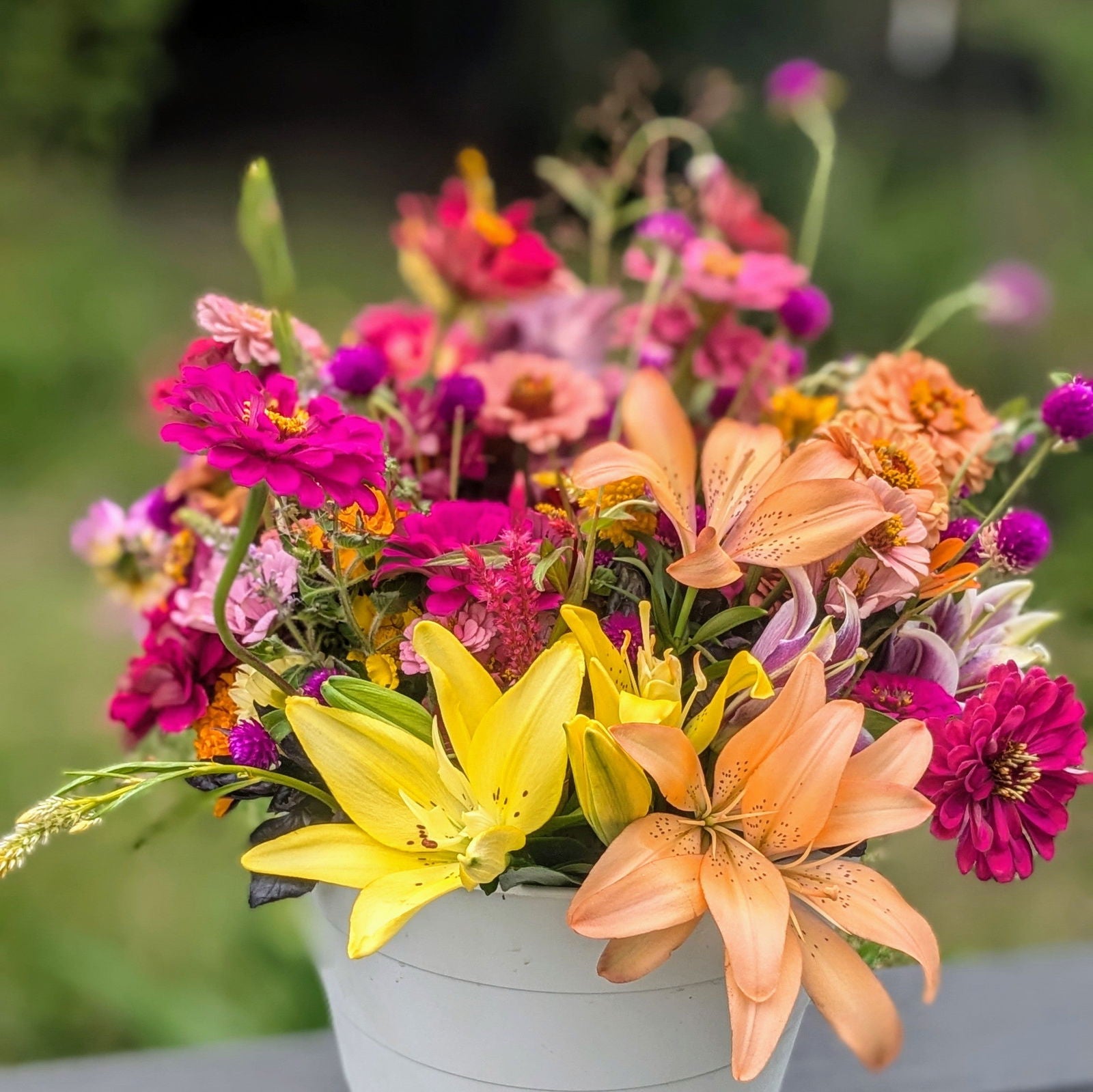 Photograph of a vibrant summer bouquet featuring lilies, zinnias, and globe amaranth in a white bucket showcasing bright yellow and peach lilies along with fuchsia and coral zinnias
