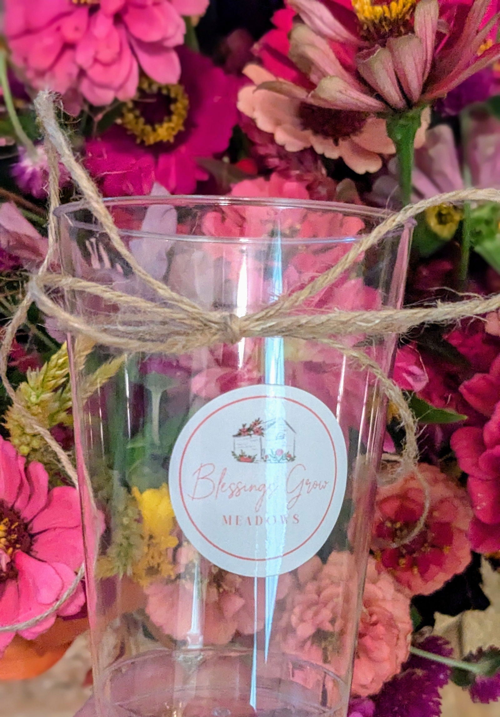 Photograph of a clear plastic cup with a Blessing Grow Meadows label surrounded by vibrant pink, purple, and yellow zinnias tied with twine.
