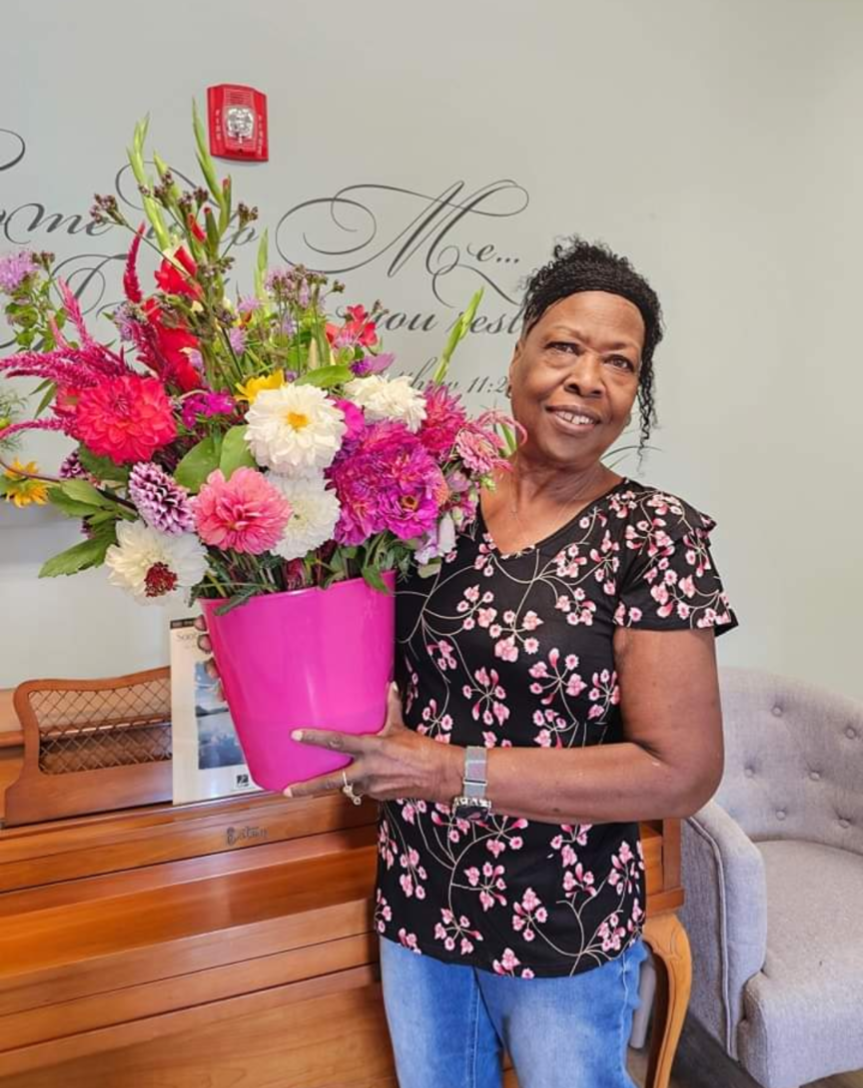 Photograph of a woman holding a vibrant pink flower arrangement featuring dahlias, gladiolus, and amaranth in a bright pink pot near a vintage wood piano and wall lettering