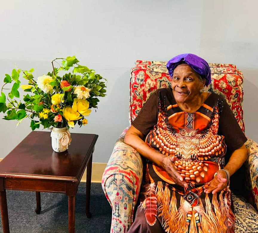 Photograph of an elderly woman in a colorful patterned dress sitting in an armchair, a vibrant floral arrangement on a nearby table, featuring yellow and white dahlias and green foliage