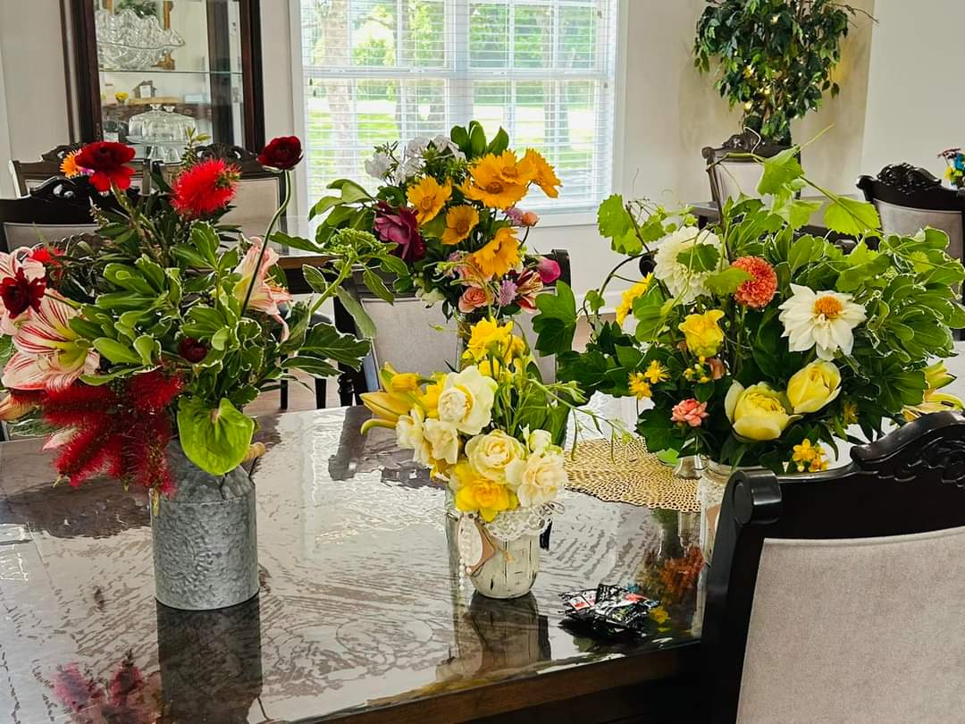 Floral arrangement featuring vibrant red, yellow, and white blooms in rustic vases on a glass-top table near a large window with a view of lush green trees