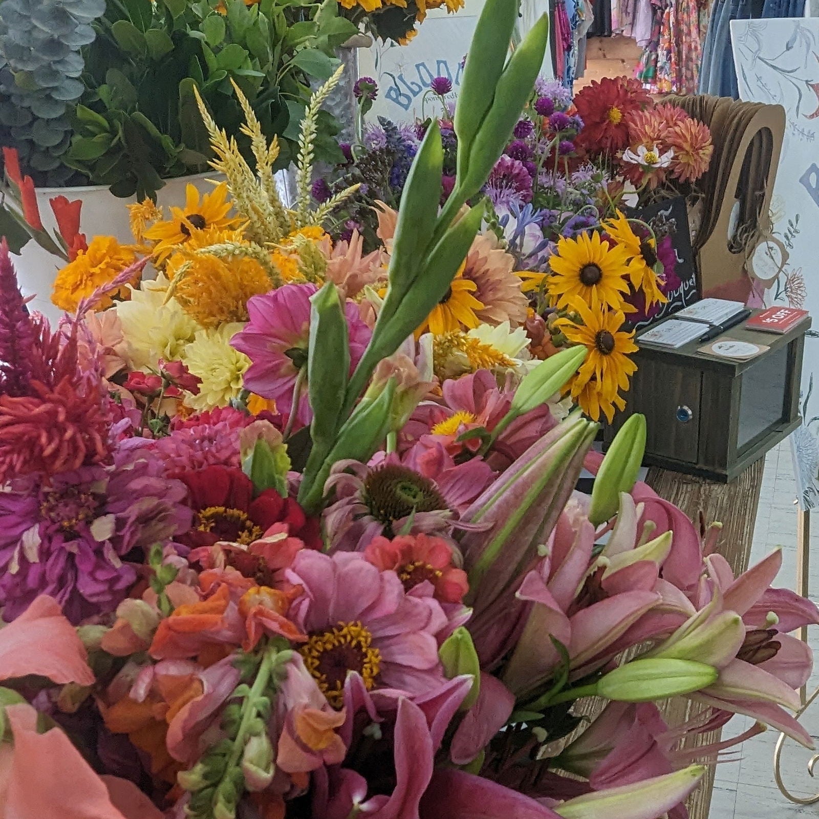 Photograph of a vibrant flower bouquet featuring lilies, zinnias, and black-eyed Susans at a flower market with a rustic wooden display case and a handwritten sign
