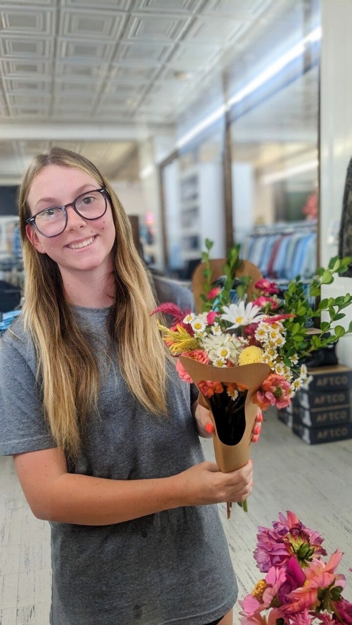 Photograph young woman holding a summer bouquet of daisies, zinnias, and orange flowers in a shop featuring high ceilings and exposed tin tiles