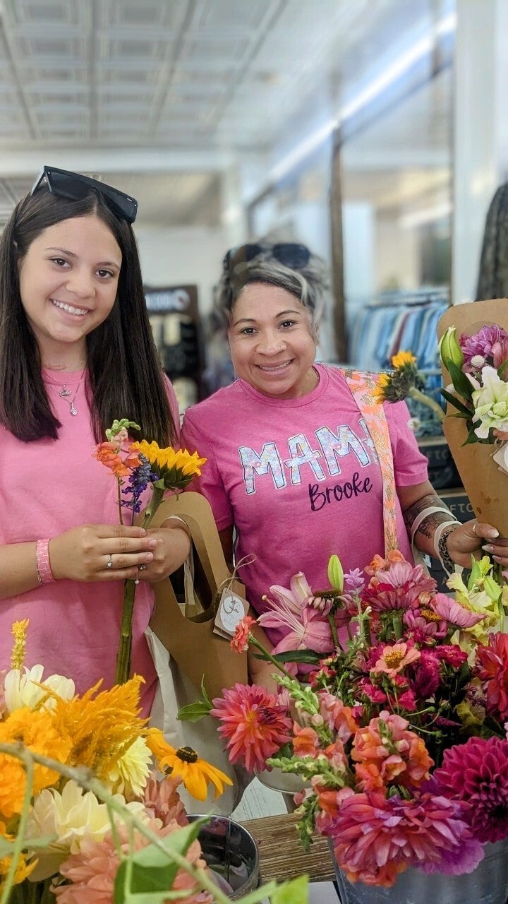 Photograph two women in pink shirts holding vibrant bouquets of sunflowers dahlias and snapdragons inside a bright shop with white ceilings