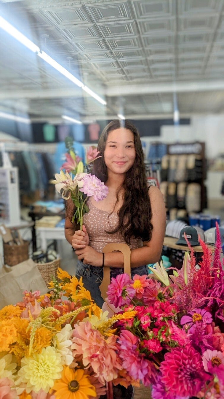 Photograph young woman holding lilies and dahlias in a flower shop with tin ceiling and colorful bouquets featuring vibrant pink and orange flowers
