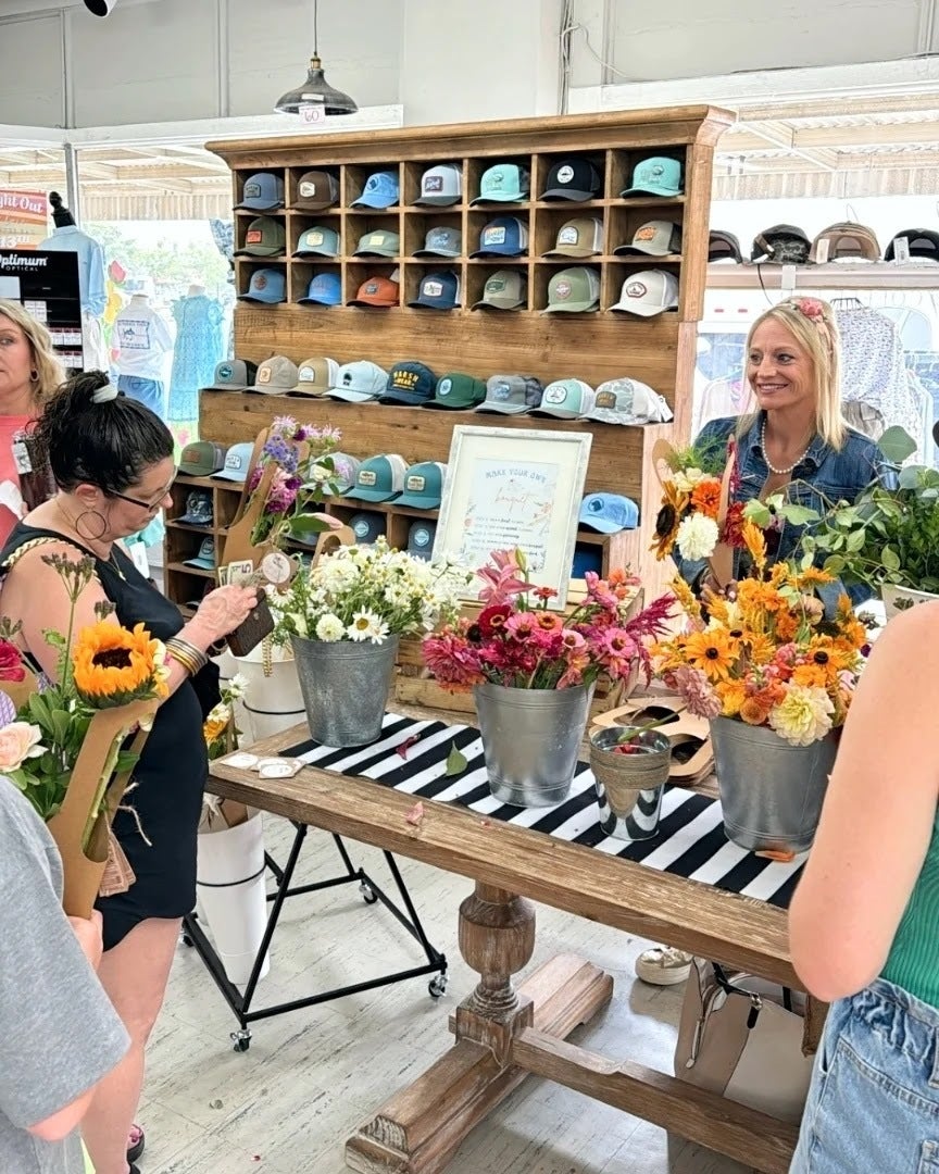 Photograph of a flower shop display featuring various colorful bouquets in galvanized buckets a rustic wooden table with black and white striped runner and a wall display of many different colored hats