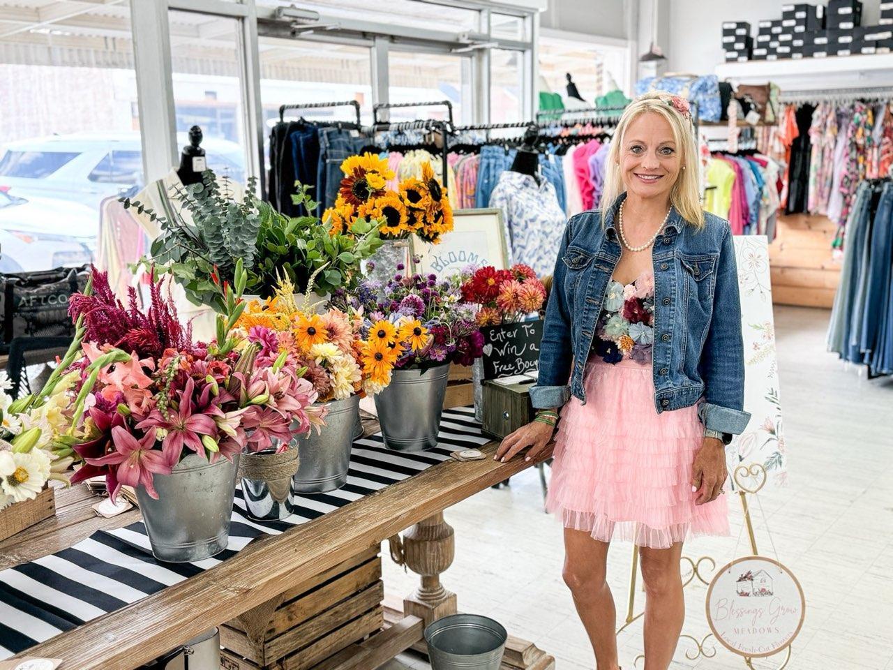 Photograph of a woman in a denim jacket and pink tulle skirt standing in Blessings Grow Meadows boutique amongst vibrant sunflowers, lilies, and dahlias in galvanized buckets displayed on a black and white striped runner
