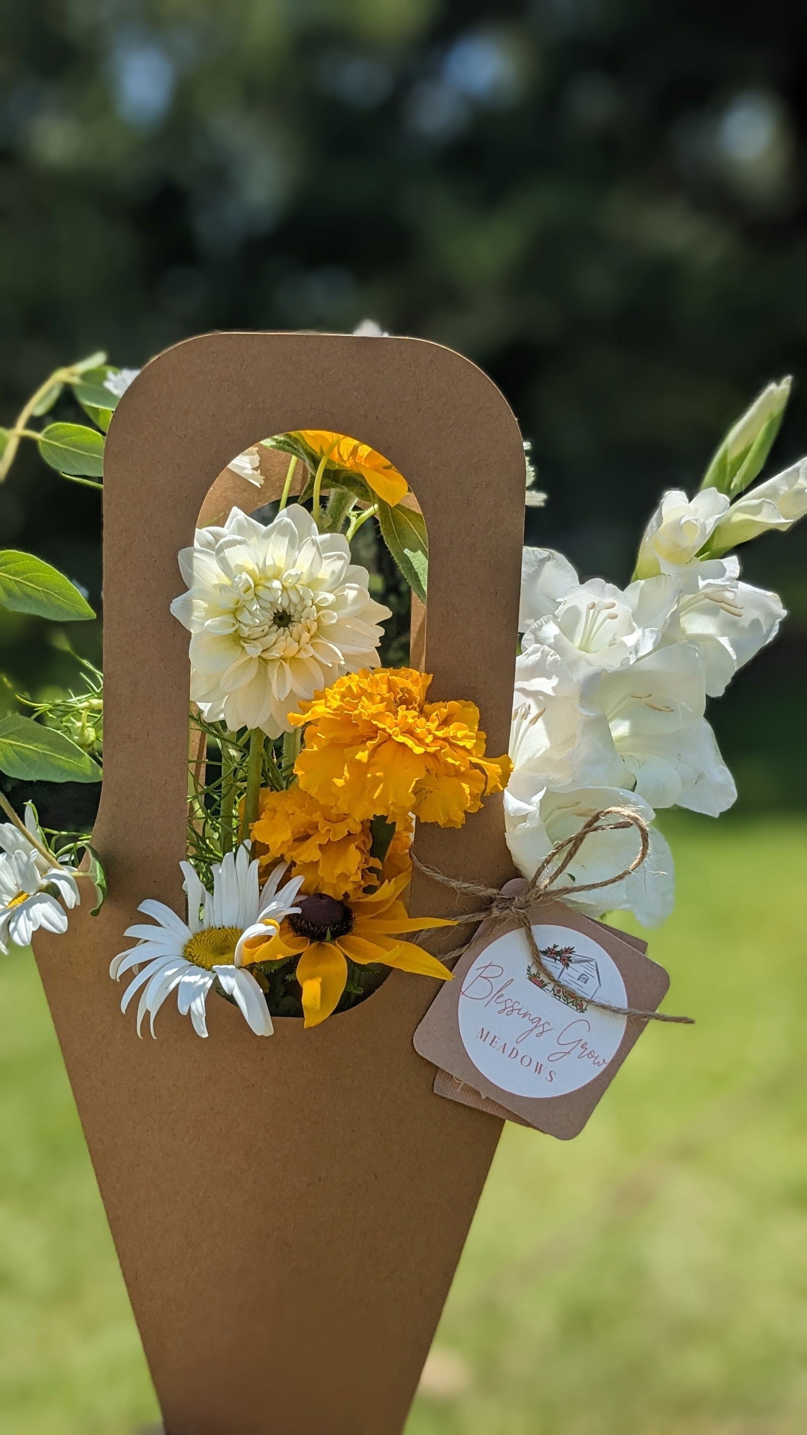 Blessings Grow Meadows floral arrangement featuring white dahlias, bright orange marigolds, and cheerful daisies in a rustic brown paper carrier
