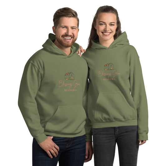 Apparel graphic design featuring a couple modeling olive green hoodies with a Blessings Grow Meadows logo depicting a charming cottage adorned with peach-colored flowers and greenery against a muted green backdrop
