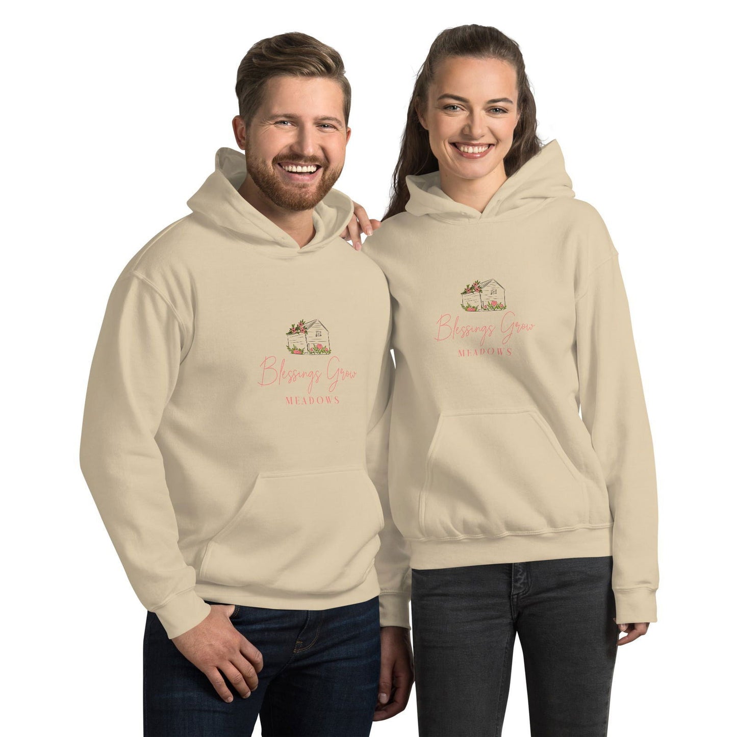 Beige hoodies featuring a Blessings Grow Meadows logo with a charming house illustration, pink flowers, and script lettering against a neutral background.
