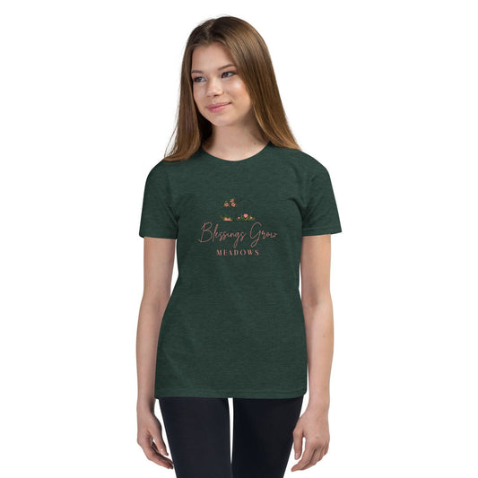 A model wearing a dark green Bella + Canvas t-shirt featuring the Blessings Grow Meadows logo with pink script and delicate floral illustrations in shades of green and pink.
