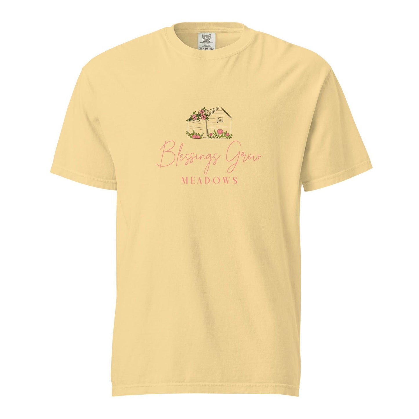 Comfort Colors t-shirt featuring a pale yellow design of a charming house adorned with pink flowers nestled in a meadow with the text Blessings Grow Meadows