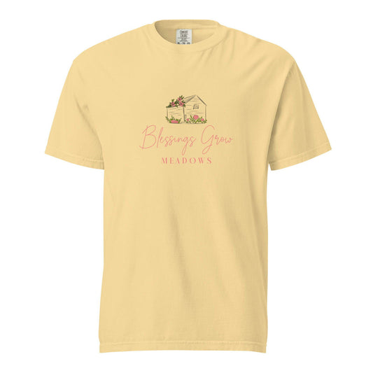 Comfort Colors t-shirt featuring a pale yellow design of a charming house adorned with pink flowers nestled in a meadow with the text Blessings Grow Meadows