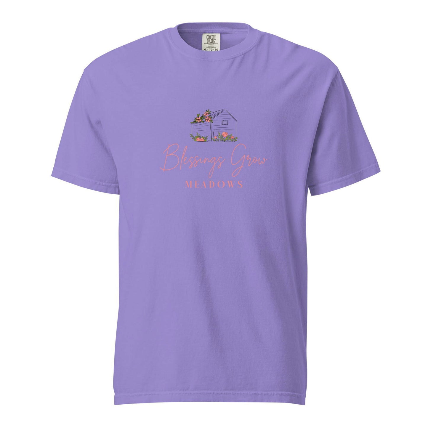 Comfort Colors t-shirt design featuring a lavender shirt with a charming illustration of a small house adorned with pink flowers and a wheelbarrow, set in a meadow with the words Blessings Grow Meadows in coral script.
