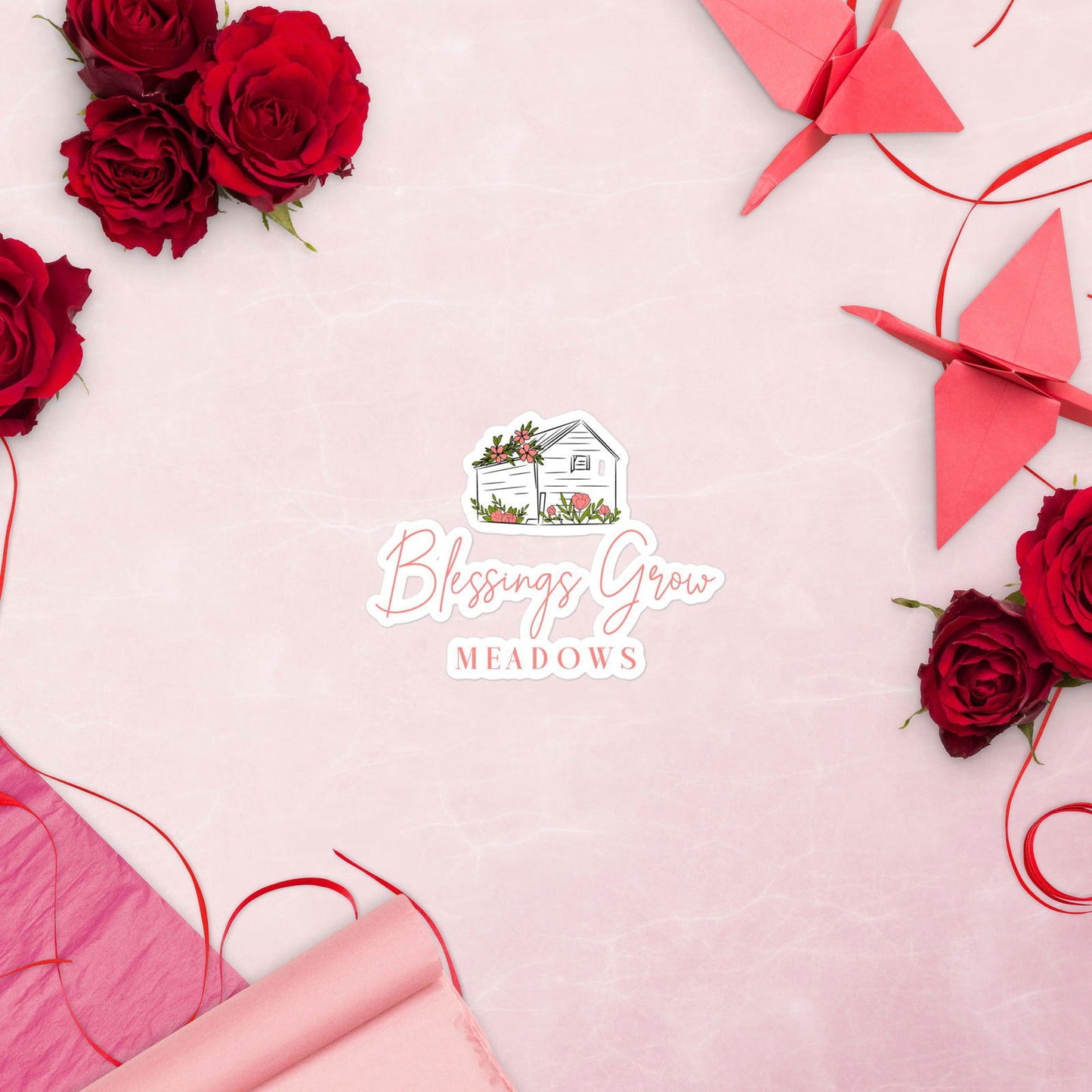 Digital illustration Blessings Grow Meadows logo featuring a charming cottage amidst pink flowers, vibrant red roses, and origami cranes on a pink background
