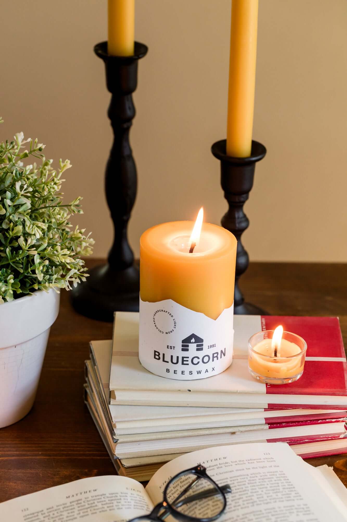Bluecorn Candles pure beeswax tea light candles in glass holders on books, with glasses and natural sunlight in a cozy home setting.