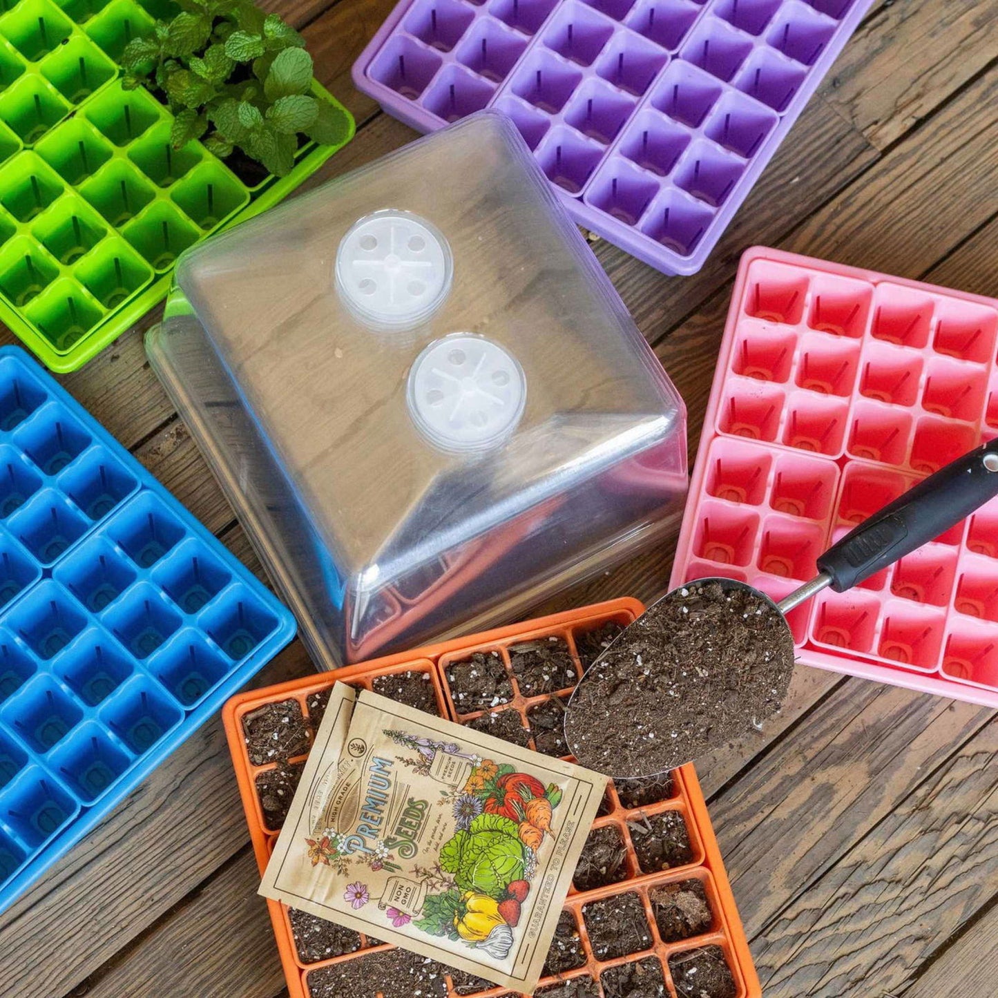 The Bootstrap Farmer 1010 Seed Starter Kit for Backyard Gardeners features colorful plastic trays, a clear humidity dome, a garden trowel with soil, and labeled “Heirloom Seeds”—perfect for starting seeds at home. Some trays hold plants or soil.