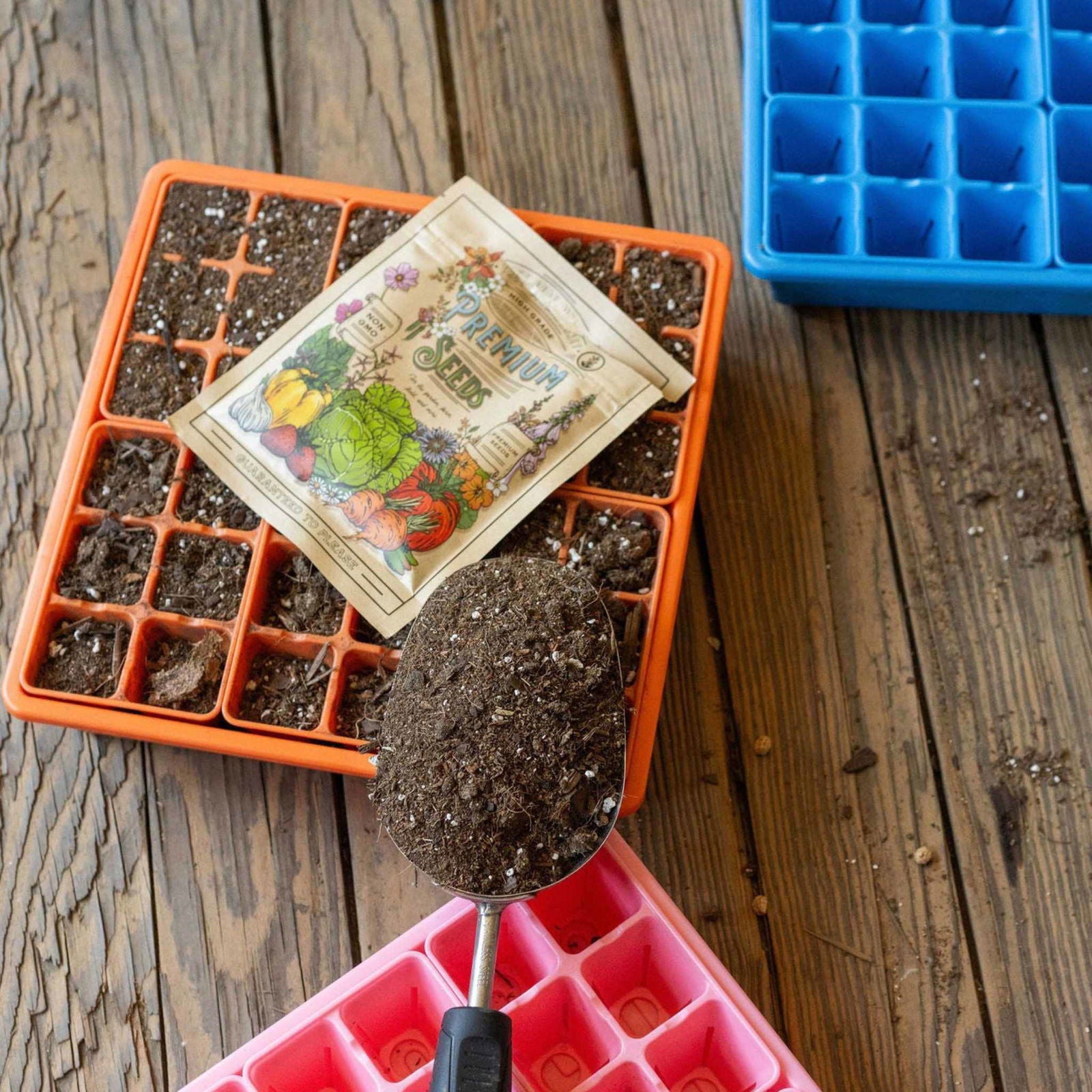 A spoon fills the Bootstrap Farmer 1010 Seed Starter Kit's colorful seedling trays with soil on a rustic table, beside a vintage vegetable seed packet—perfect for backyard gardeners ready to plant.