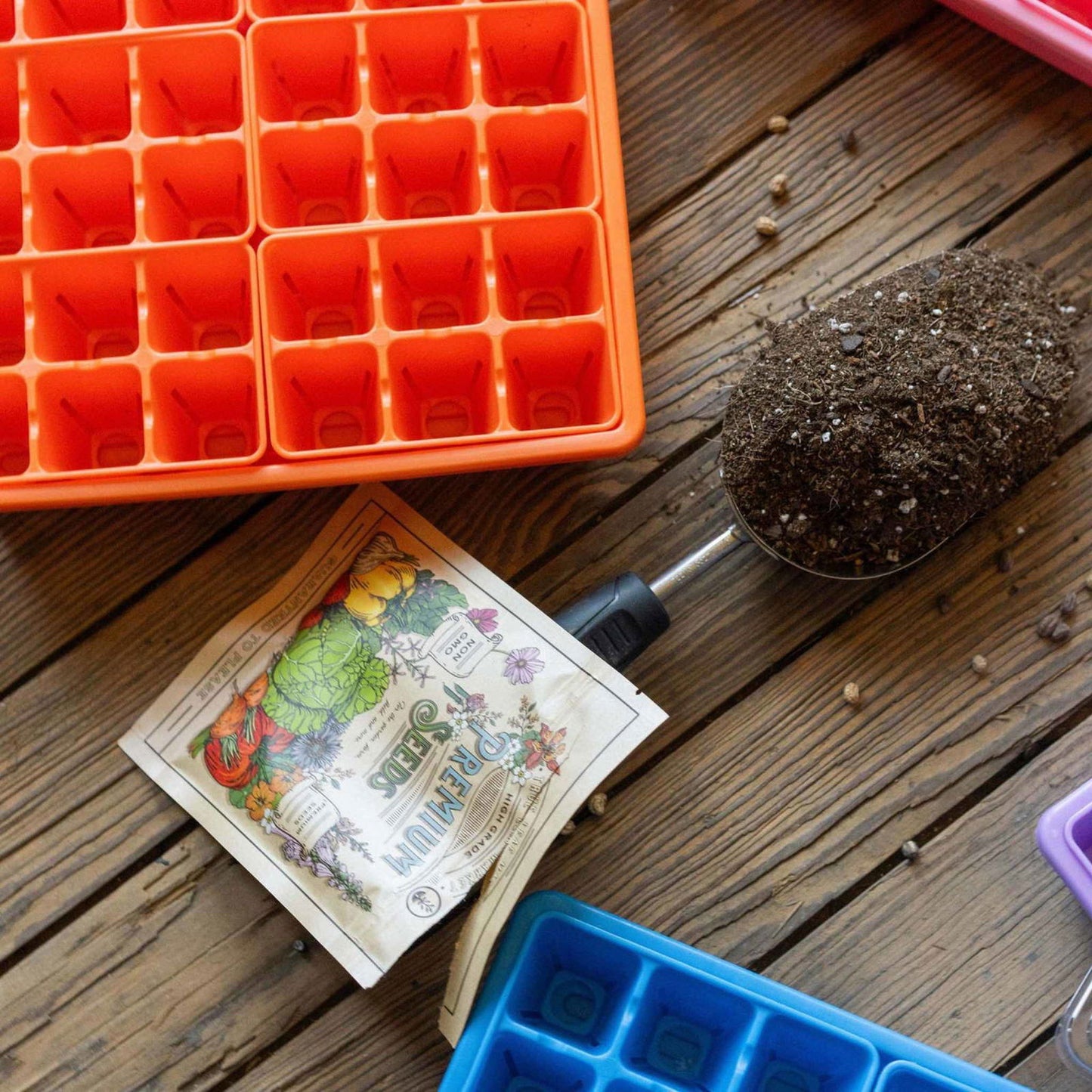 The Bootstrap Farmer 1010 Seed Starter Kit for Backyard Gardeners includes a trowel, soil, seed packets, and colorful plastic trays, all arranged on a wooden table and ready for planting.