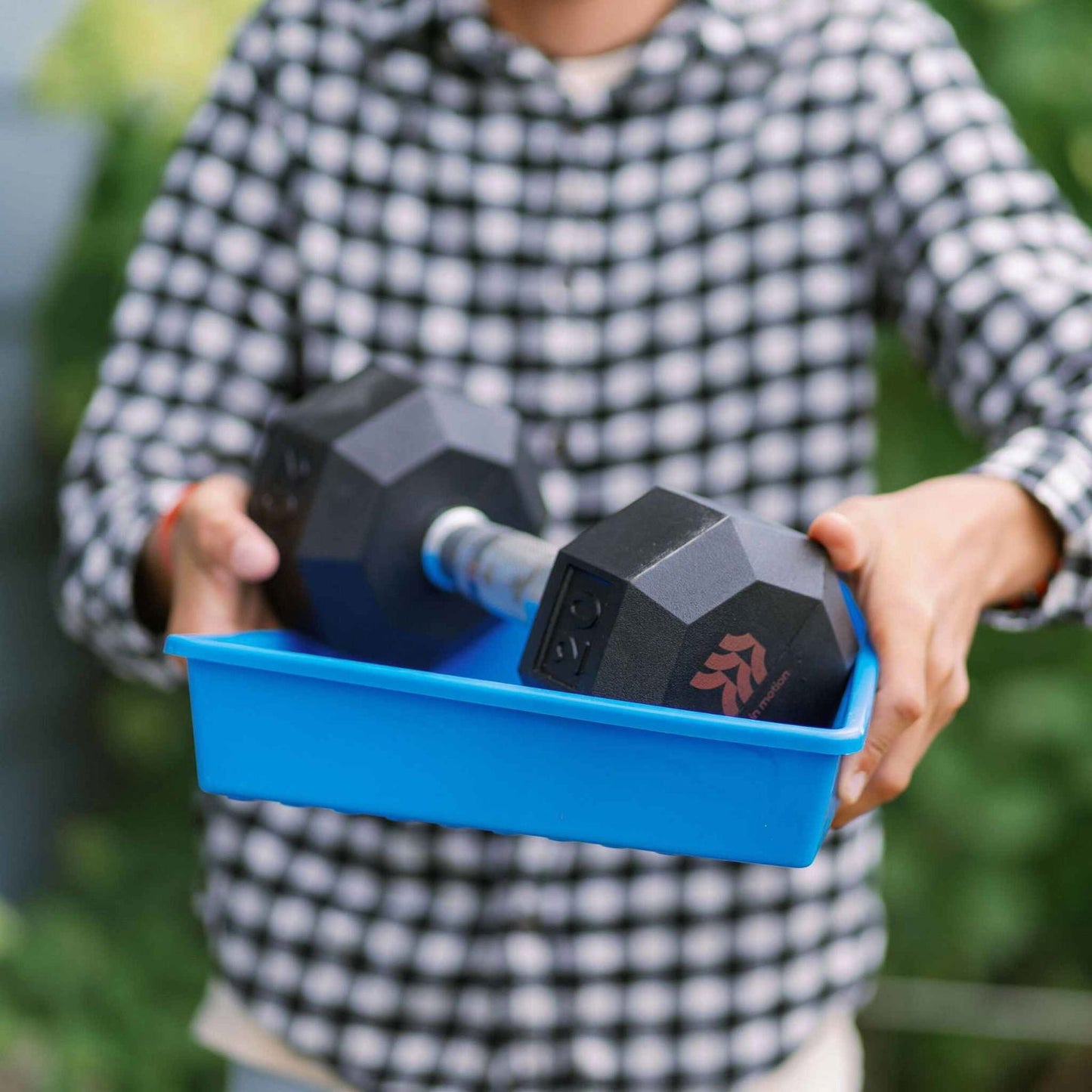 A person in a plaid shirt holds the Bootstrap Farmer 1010 Seed Starter Kit tray, featuring a black hexagonal dumbbell. Blurred green foliage behind makes it ideal for backyard gardeners.