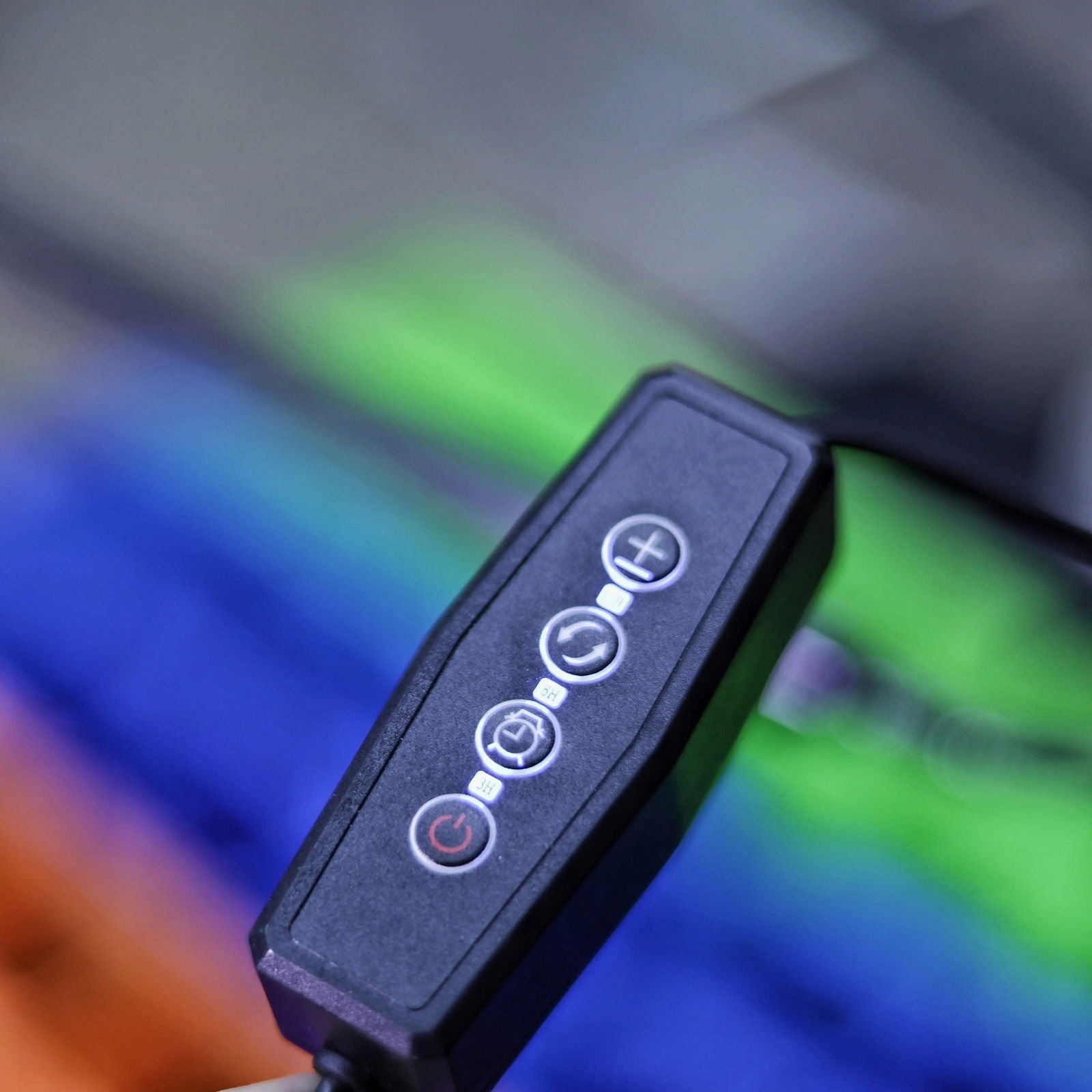 Close-up photograph of a black control panel with plus, minus, timer, and power buttons against a blurred background of bright green, blue, and orange hues.
