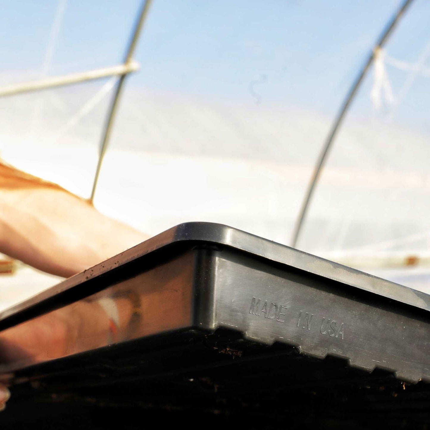 Plastic tray Made in USA in a greenhouse showing a person's arm and the metal frame of the structure with a bright sunny sky in the background