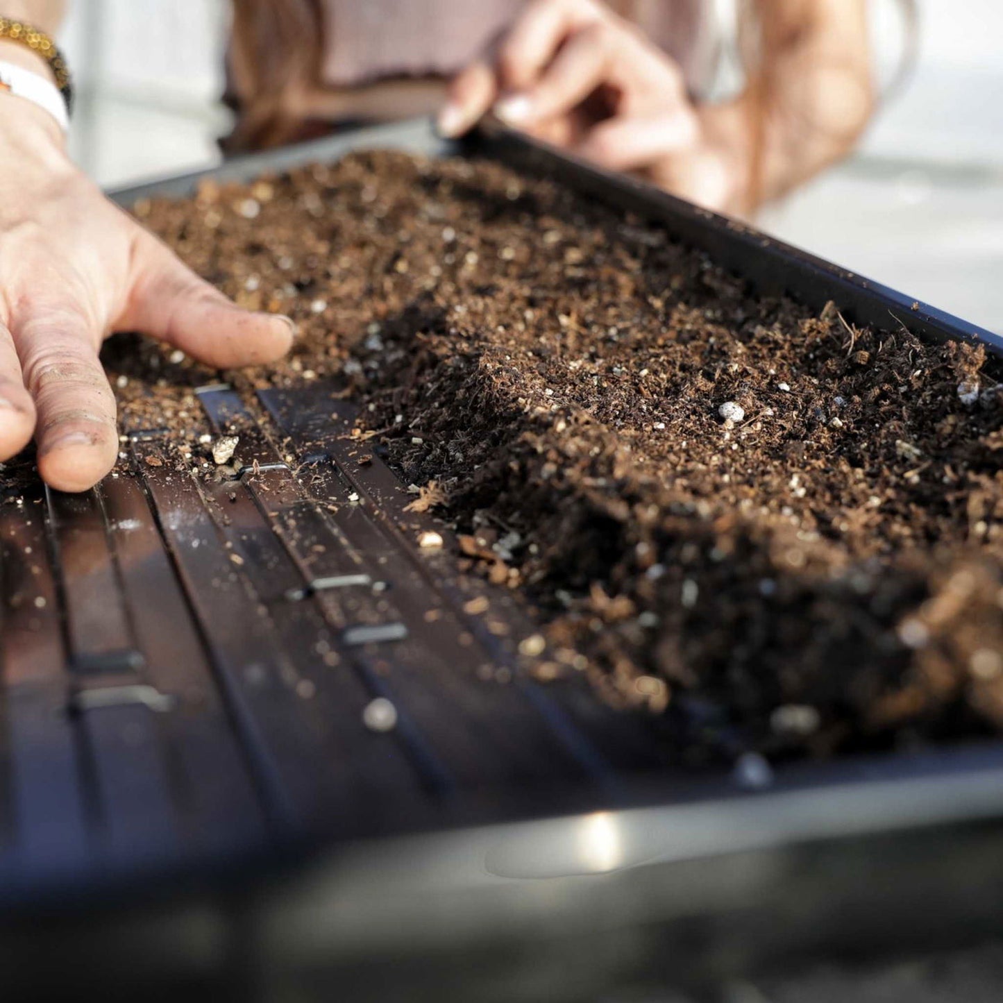 Photograph of hands preparing dark rich soil in a black plastic planter box with visible drainage slots and small white fertilizer pellets