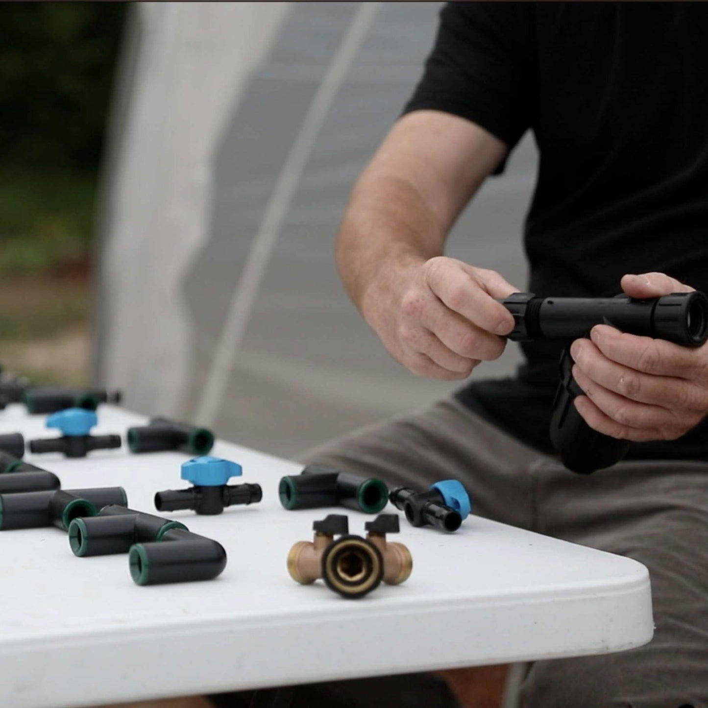 Photograph showing a person assembling black and blue irrigation parts on a white table outdoors near a greenhouse featuring a brass valve and green accents.
