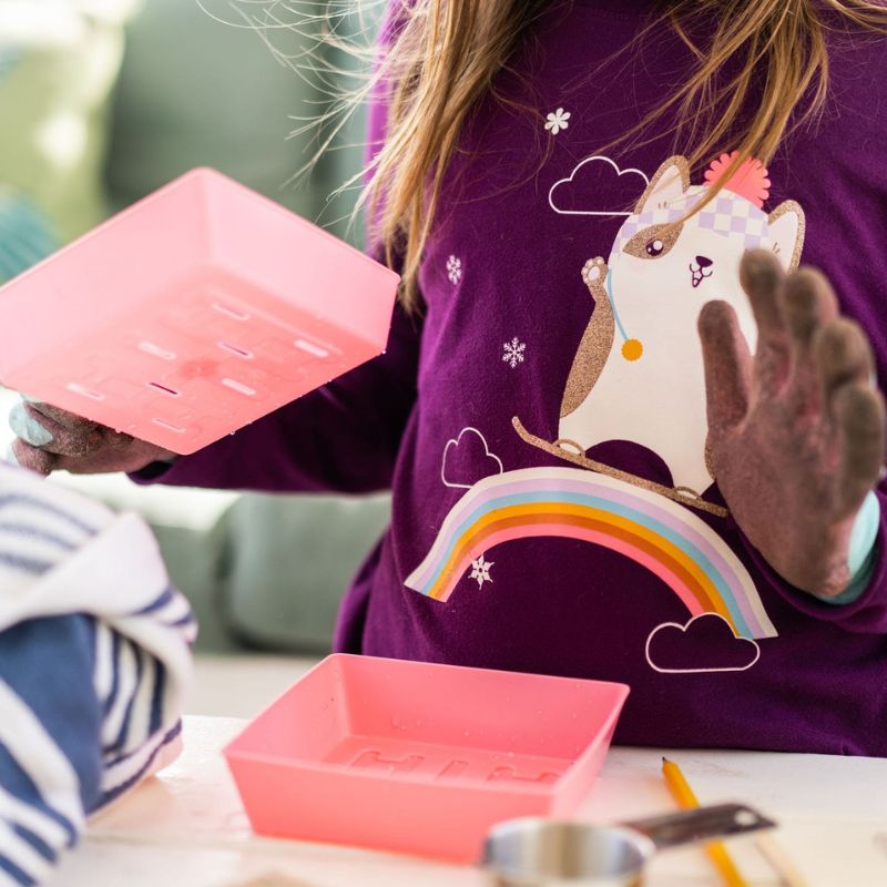 Photograph of a child wearing a purple long-sleeved shirt featuring a rainbow and a snowboarding raccoon holding two pink plastic trays with a colorful design and muddy hands.
