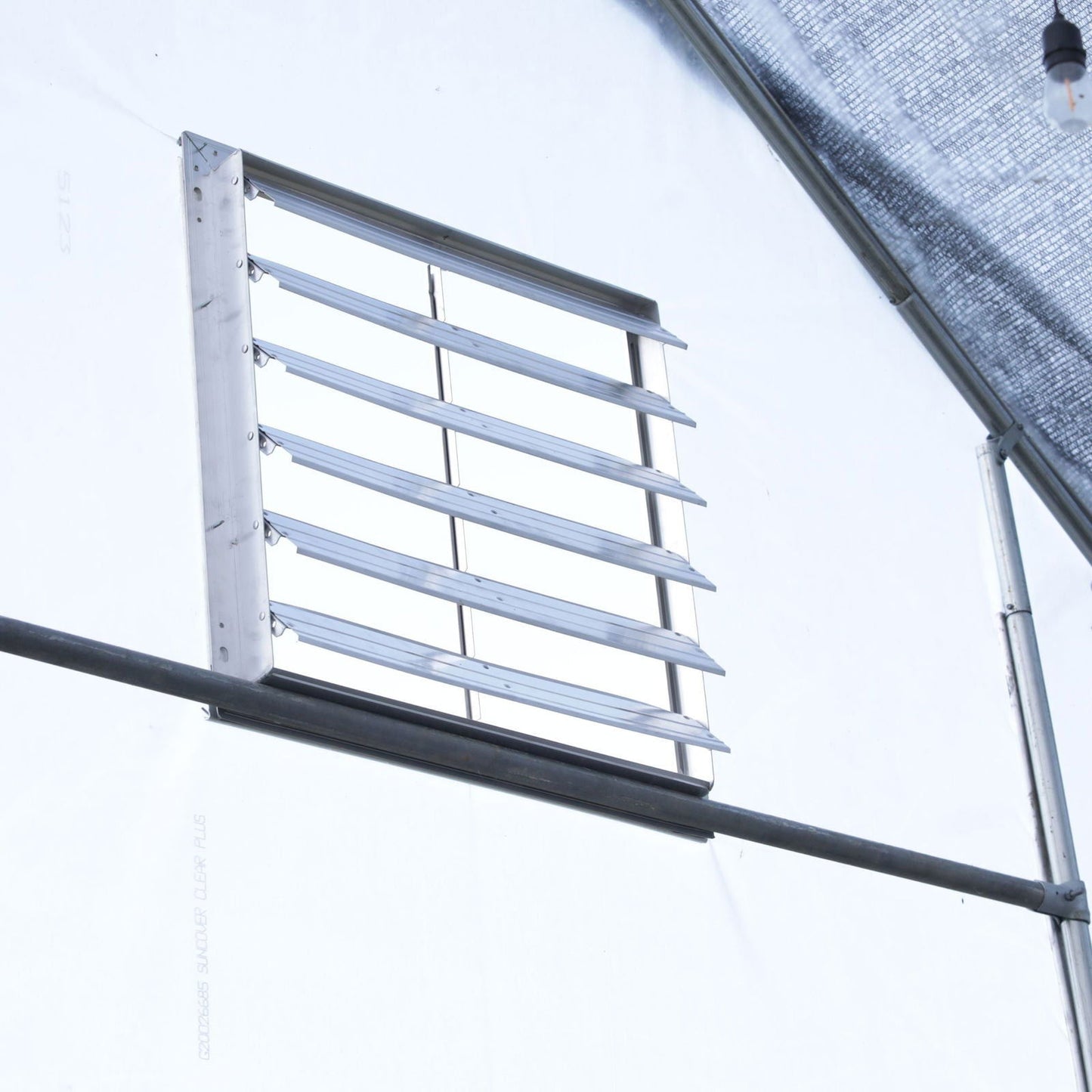A Bootstrap Farmer Greenhouse Aluminum Intake Shutter with angled slats for airflow is installed on a white wall. Nearby, a black horizontal pipe runs along the structure, with a translucent cover partially visible above.