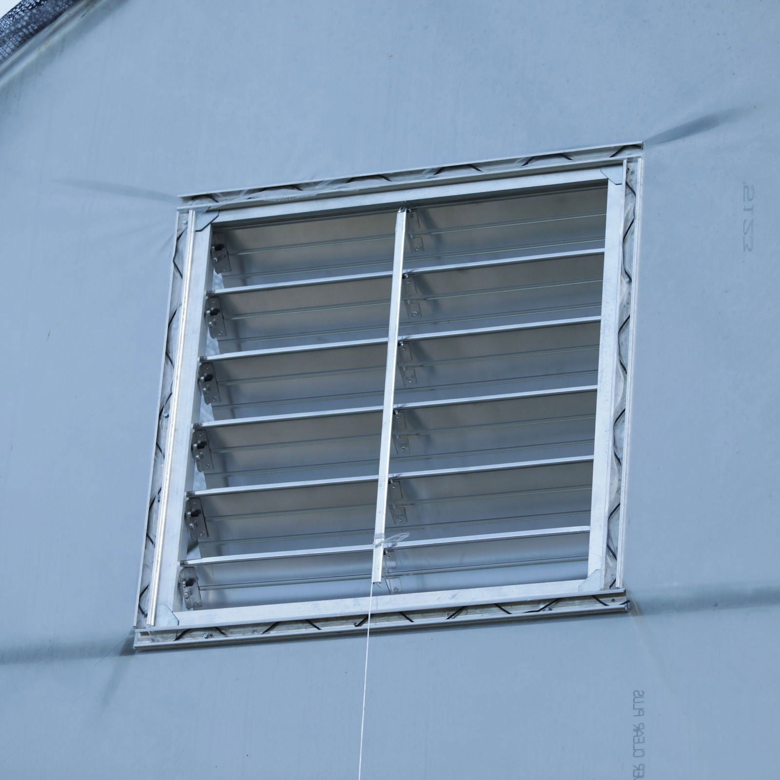 The Bootstrap Farmer Greenhouse Aluminum Intake Shutter is mounted on a smooth, light blue wall. Its horizontal slats enhance ventilation, while a thin cord hangs vertically from the center for easy operation.
