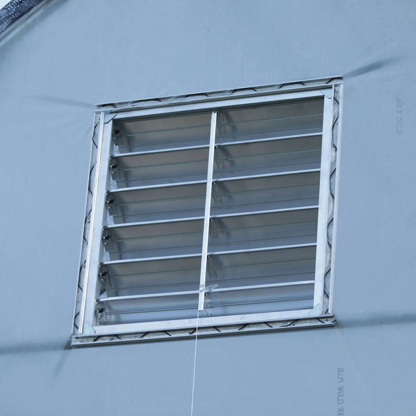 A close-up of the Bootstrap Farmer Greenhouse Aluminum Intake Shutter with horizontal slats, set in a light blue wall. A white cord hangs vertically, ideal for greenhouse ventilation.