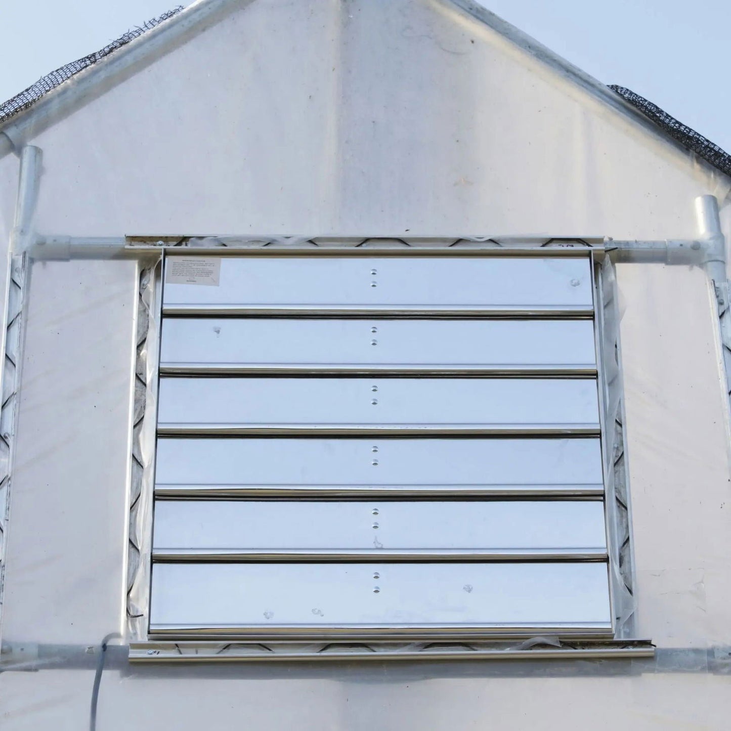 A close-up of a Bootstrap Farmer Greenhouse Shutter Mount Exhaust Fan centered in a wall, framed by translucent plastic panels and metal supports—ideal for efficient airflow in any hoop house.