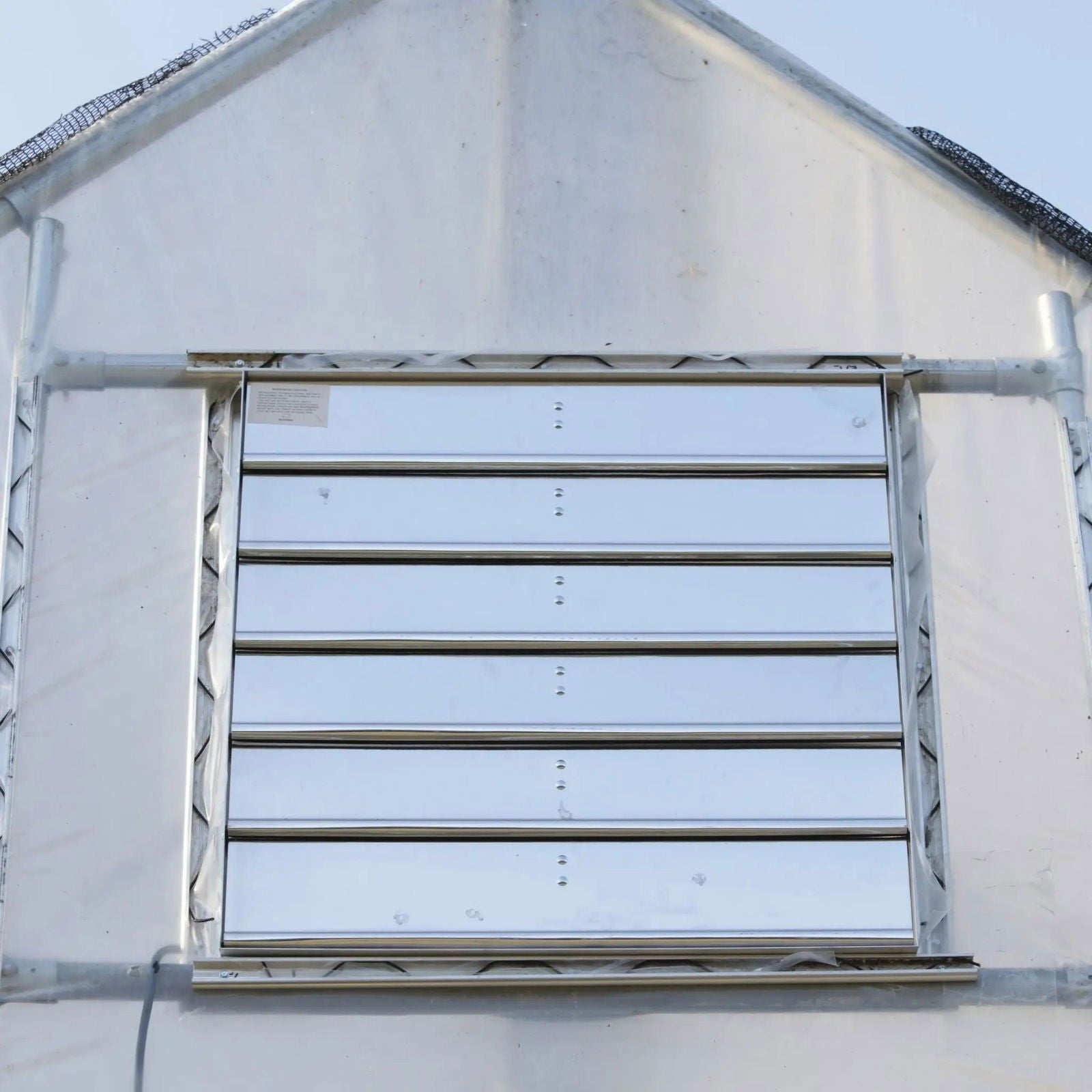 A close-up of a Bootstrap Farmer Greenhouse Shutter Mount Exhaust Fan centered in a wall, framed by translucent plastic panels and metal supports—ideal for efficient airflow in any hoop house.