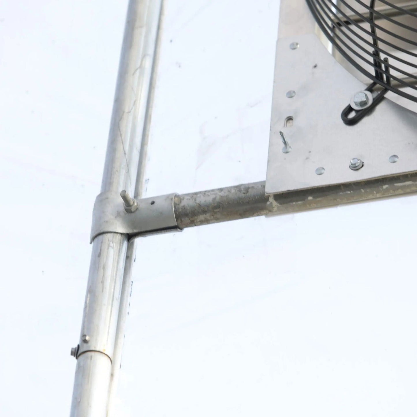 A close-up of Bootstrap Farmer’s Greenhouse Shutter Mount Exhaust Fans shows metal pipes and clamps next to a metal panel and exhaust fan grille, set against a light background—ideal for greenhouse or hoop house construction.