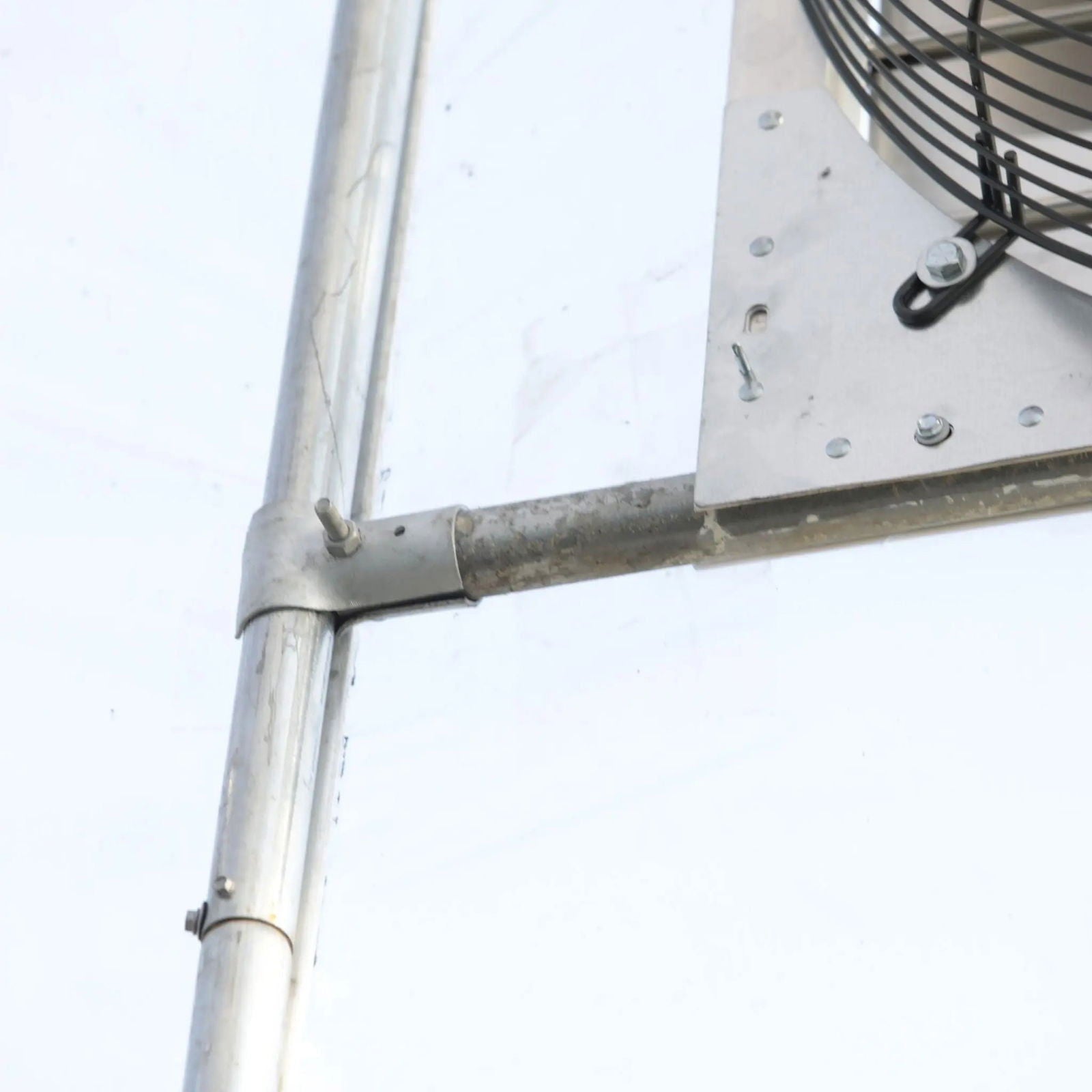 A close-up of Bootstrap Farmer’s Greenhouse Shutter Mount Exhaust Fans shows metal pipes and clamps next to a metal panel and exhaust fan grille, set against a light background—ideal for greenhouse or hoop house construction.